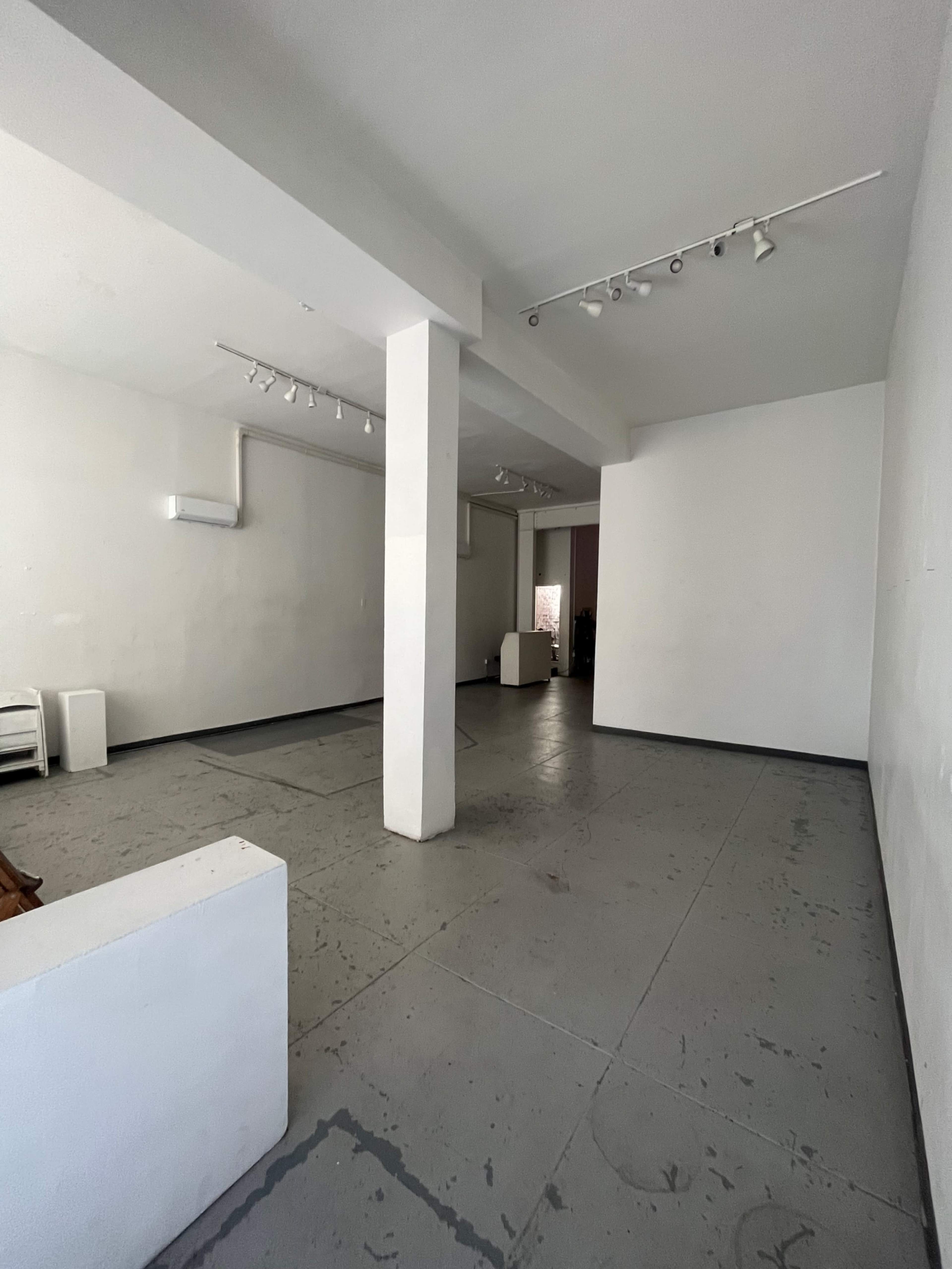 An empty, minimalist room with white walls and a concrete floor, featuring a central pillar and several pieces of white furniture against the walls.