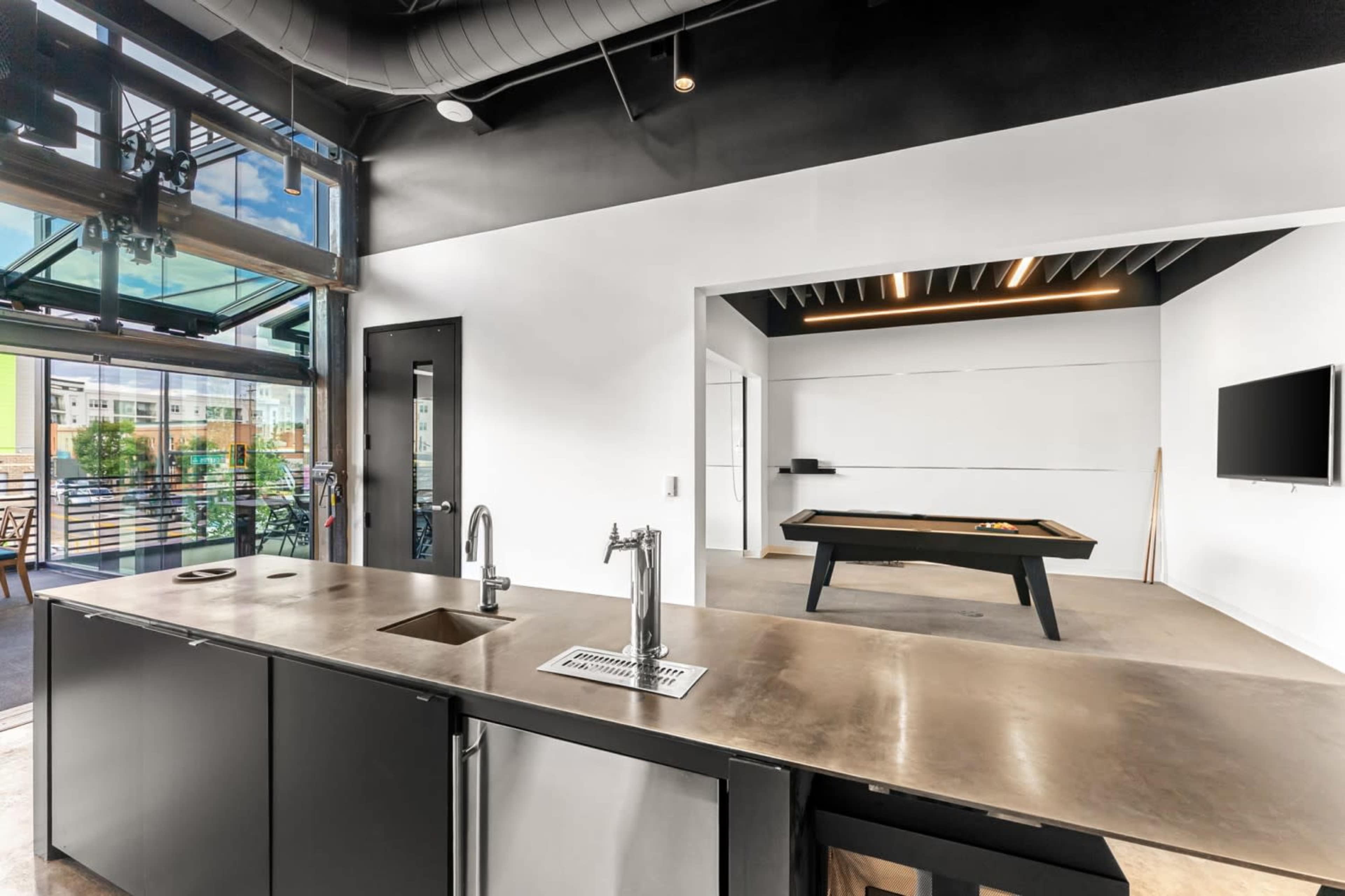 The image shows a modern open-concept office space featuring a sleek kitchen area with a metal countertop and a pool table in a well-lit room.