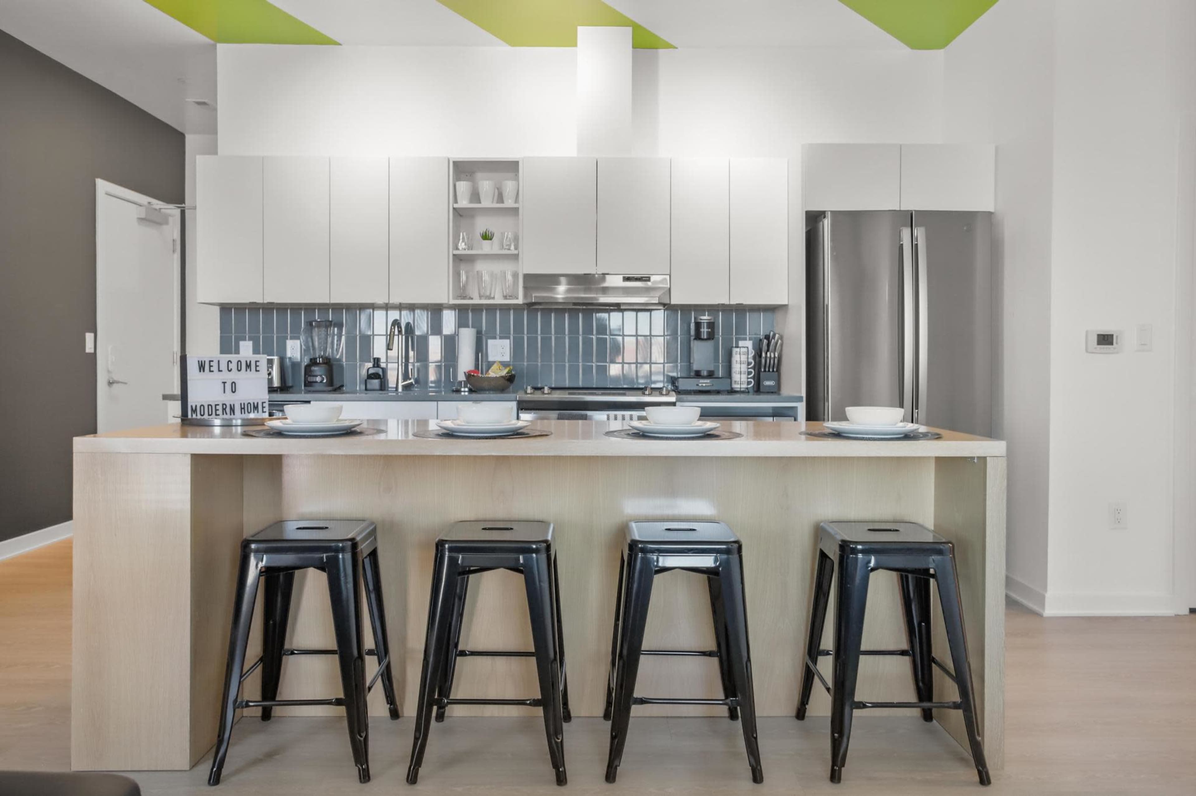 A modern kitchen features a central island with four black stools, white cabinets, and a stainless steel refrigerator.
