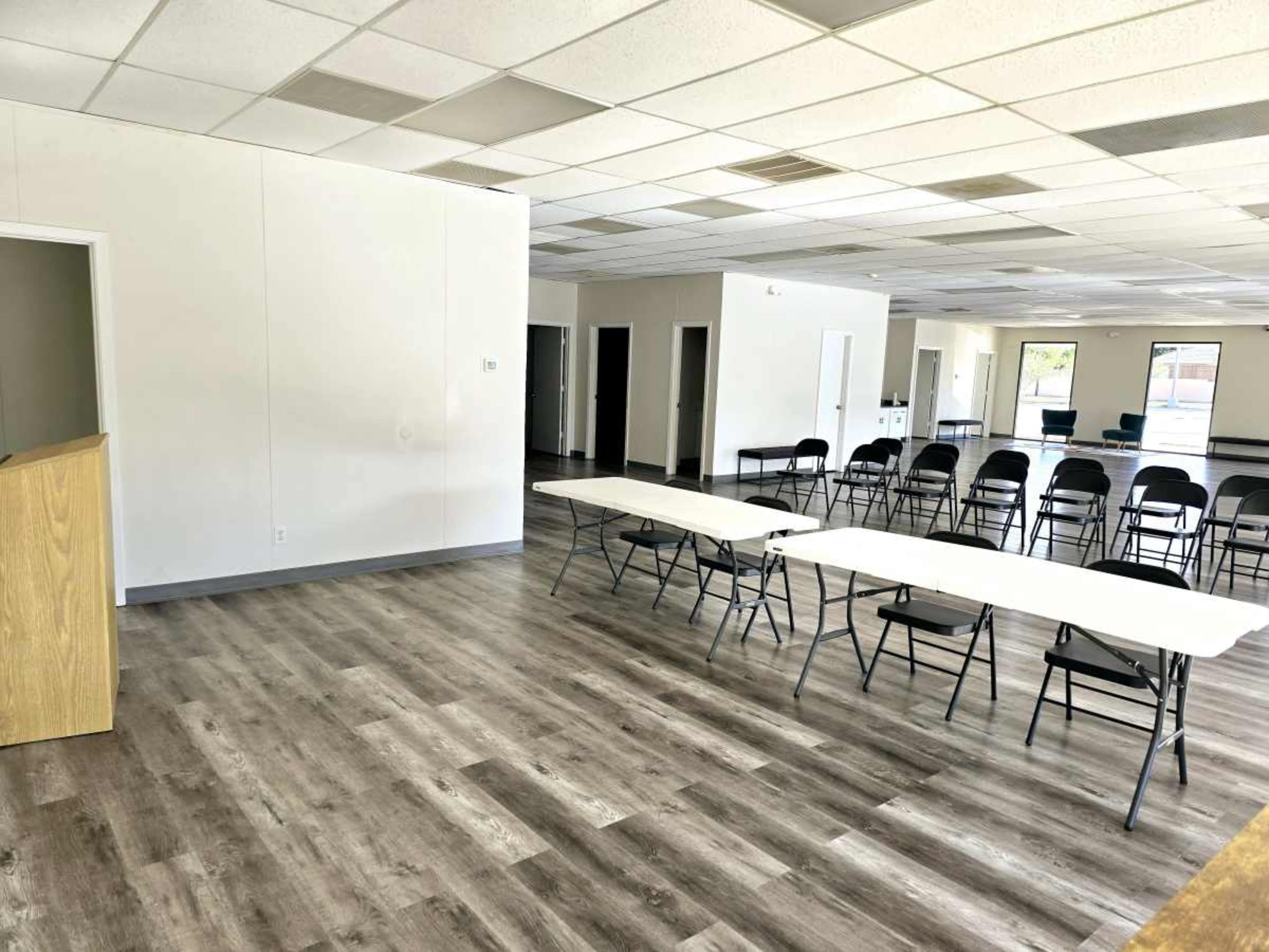 An empty room features a wooden podium, several folding tables arranged in a row, and chairs set up for potential seating, with a light-colored floor and plain walls.