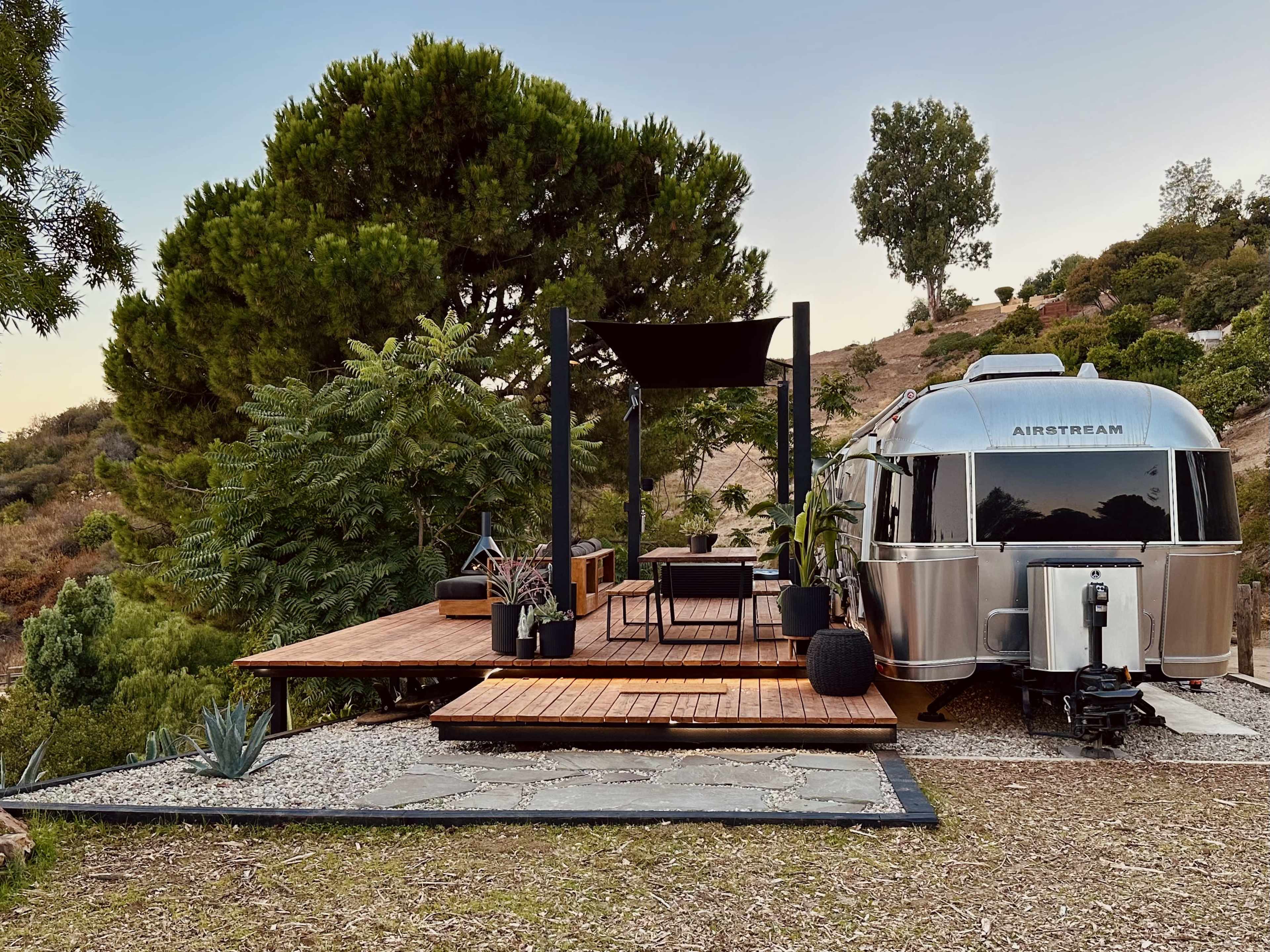 A silver Airstream trailer is parked next to a wooden deck furnished with chairs and plants, surrounded by greenery on a hillside.