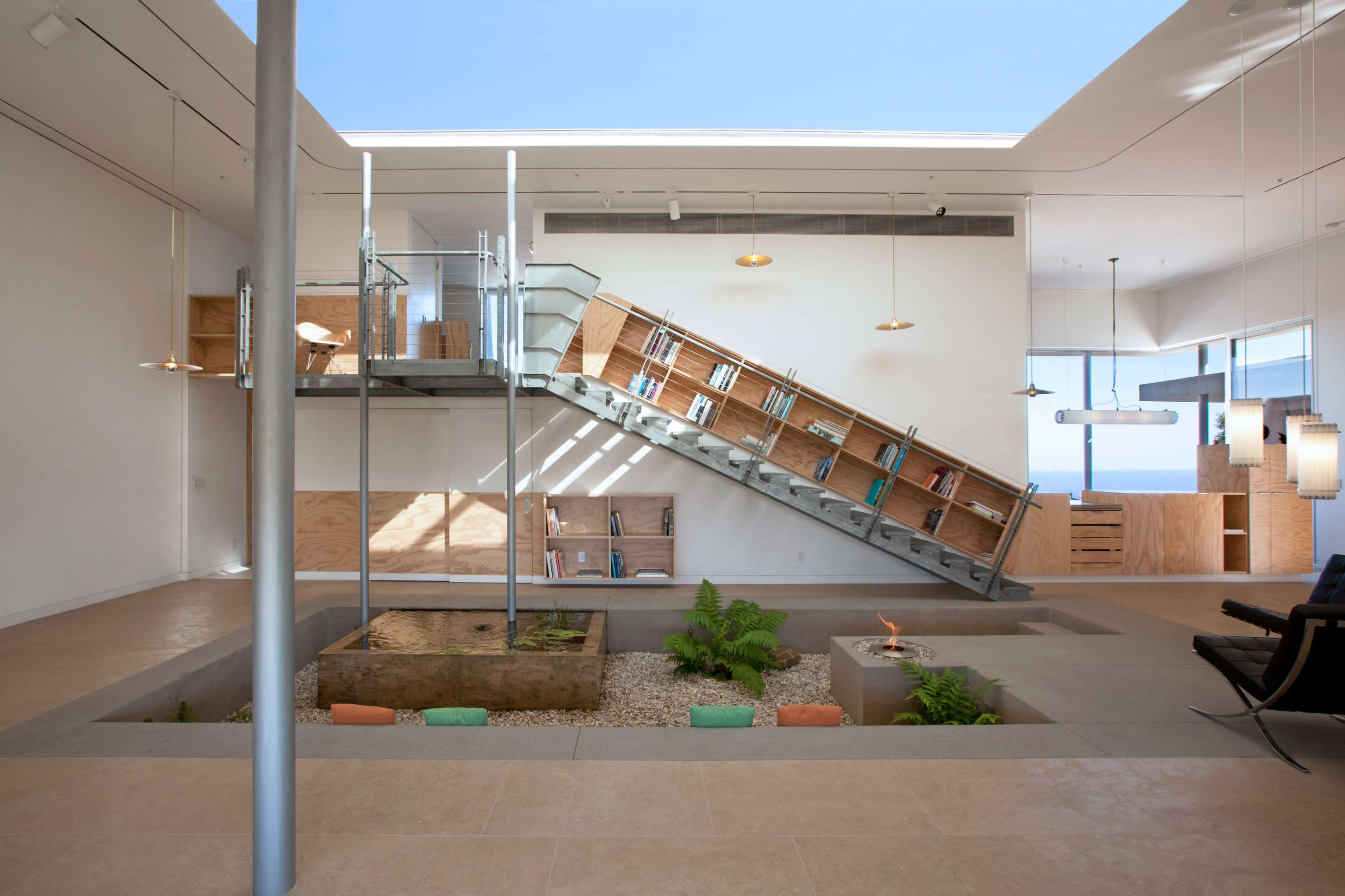 The image shows a modern interior space featuring a large staircase, an indoor garden with a water feature, and large windows that allow natural light to fill the room.