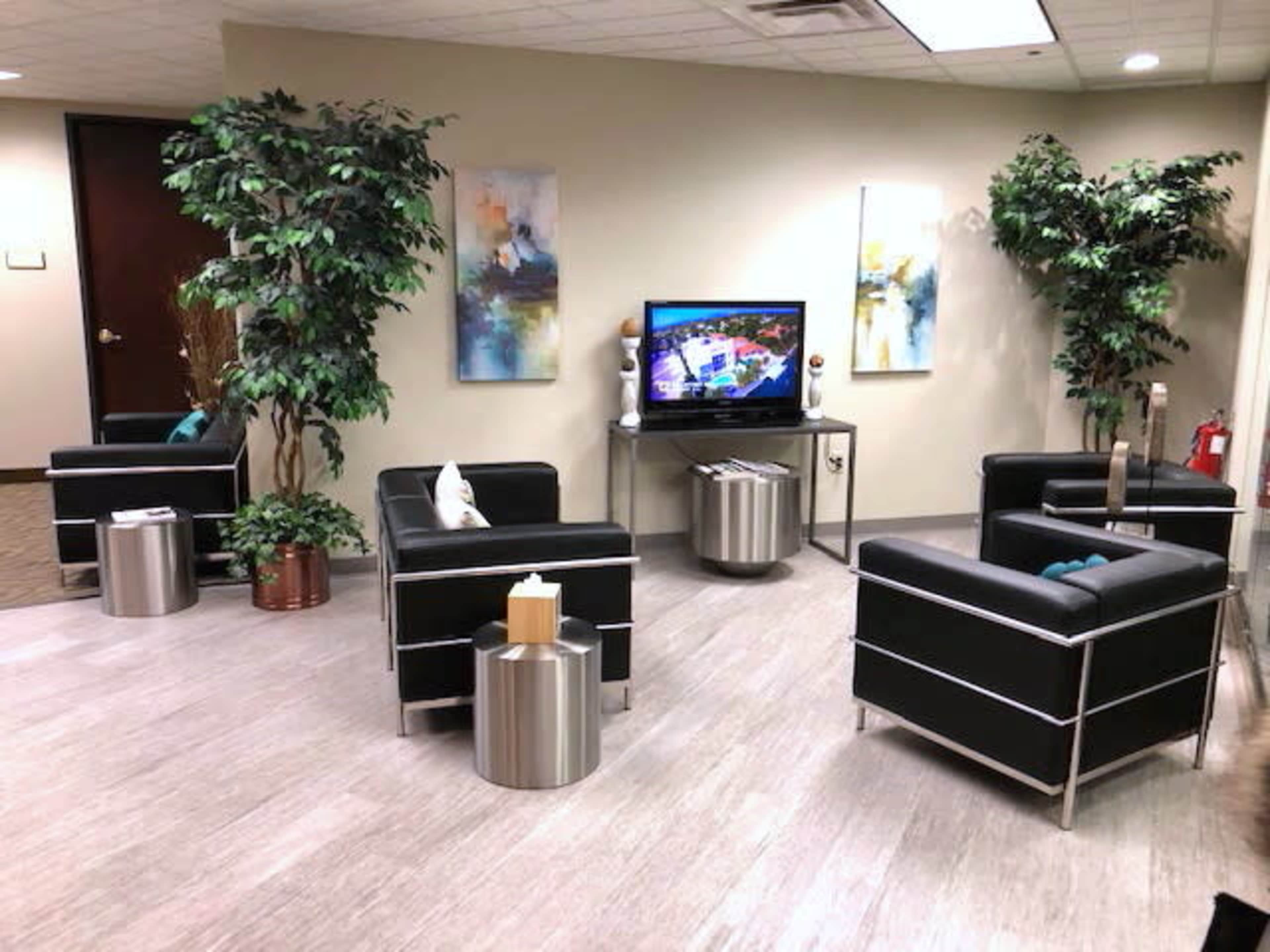 The image shows a waiting area with black chairs, small metallic tables, and decorative plants, featuring a television mounted on the wall.
