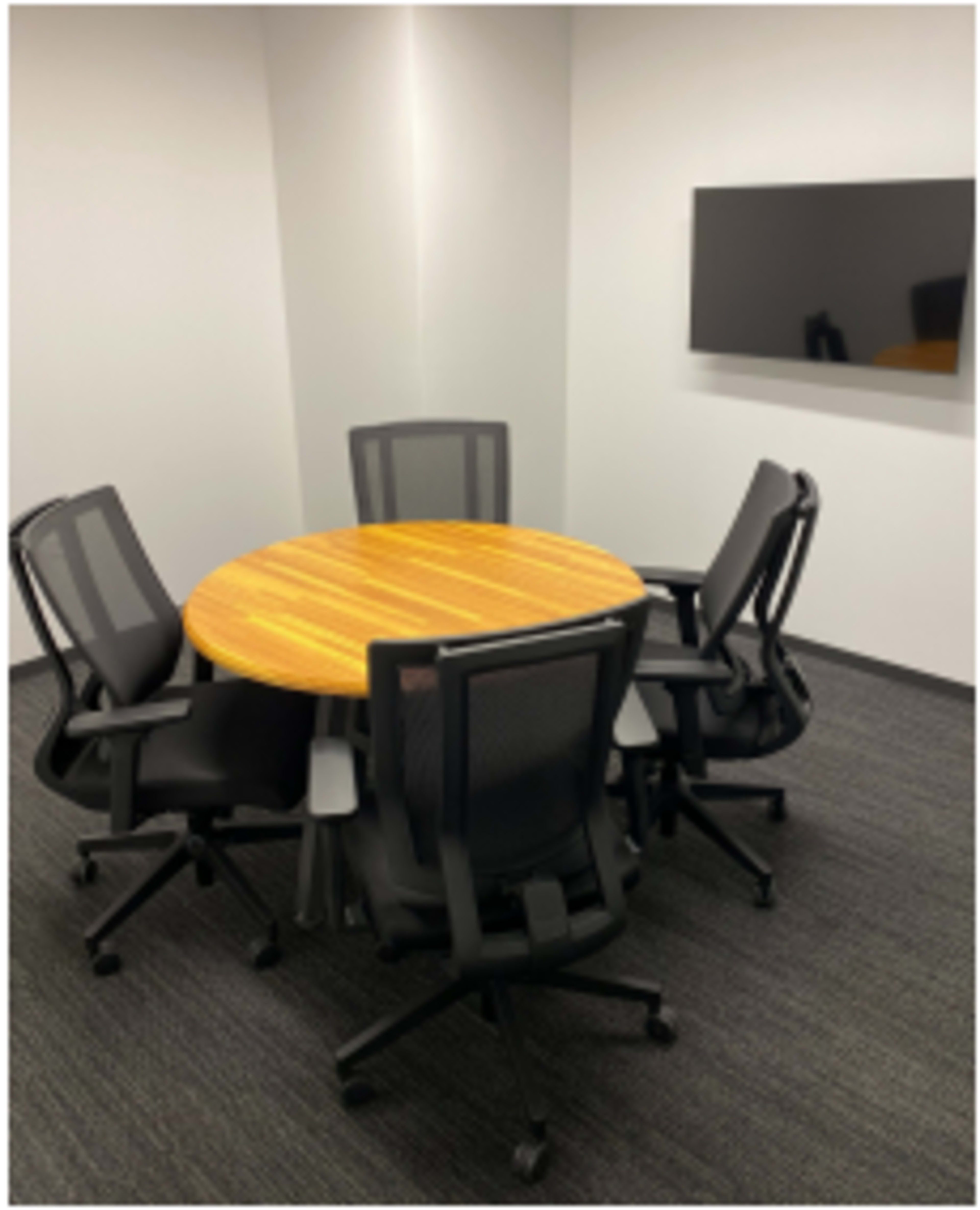 A round wooden table with five black rolling chairs is set up in a small, empty conference room.