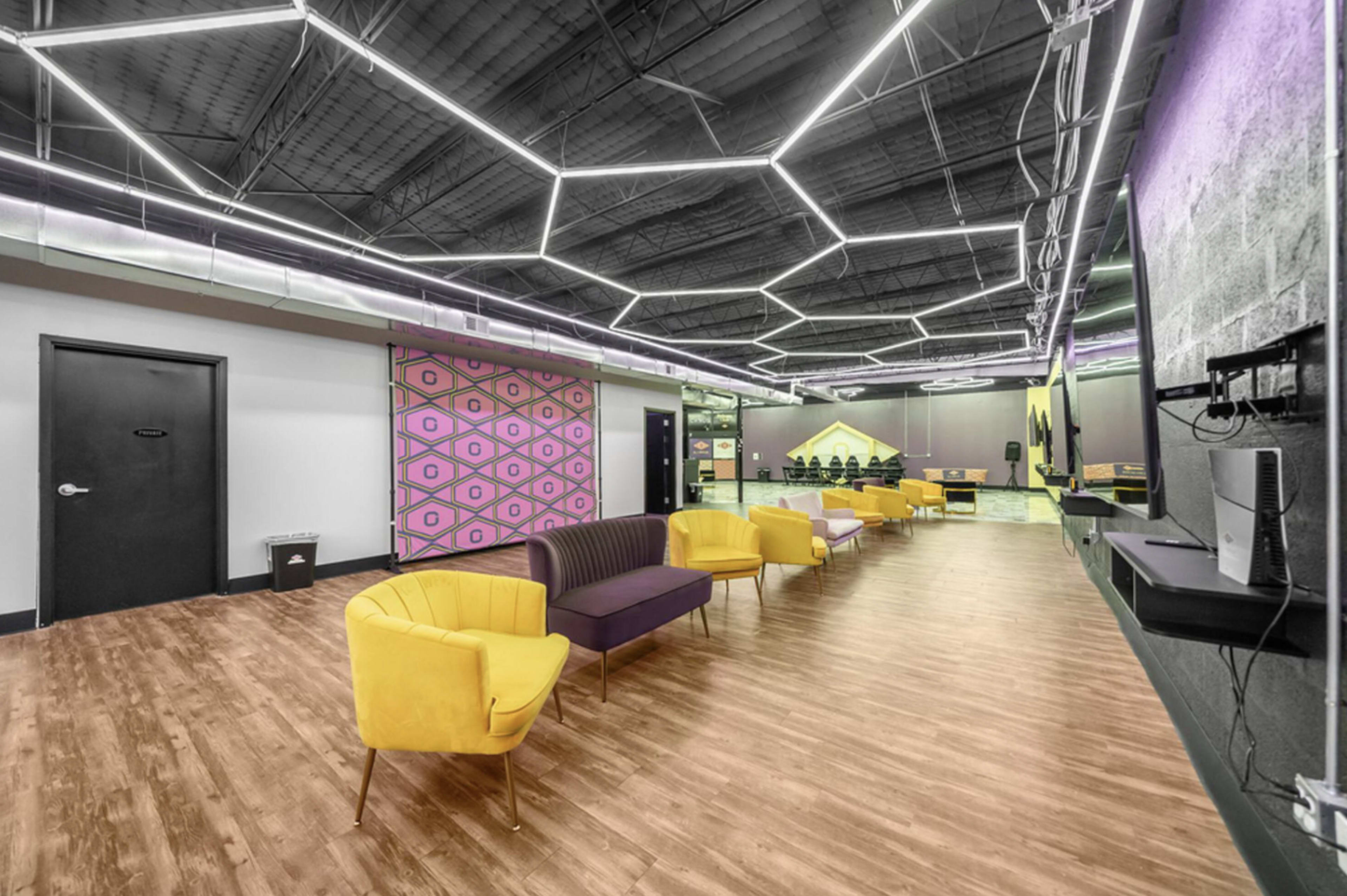 The image shows a modern interior space with yellow and purple chairs arranged along a wooden floor, featuring geometric ceiling lights and decorative wall elements.