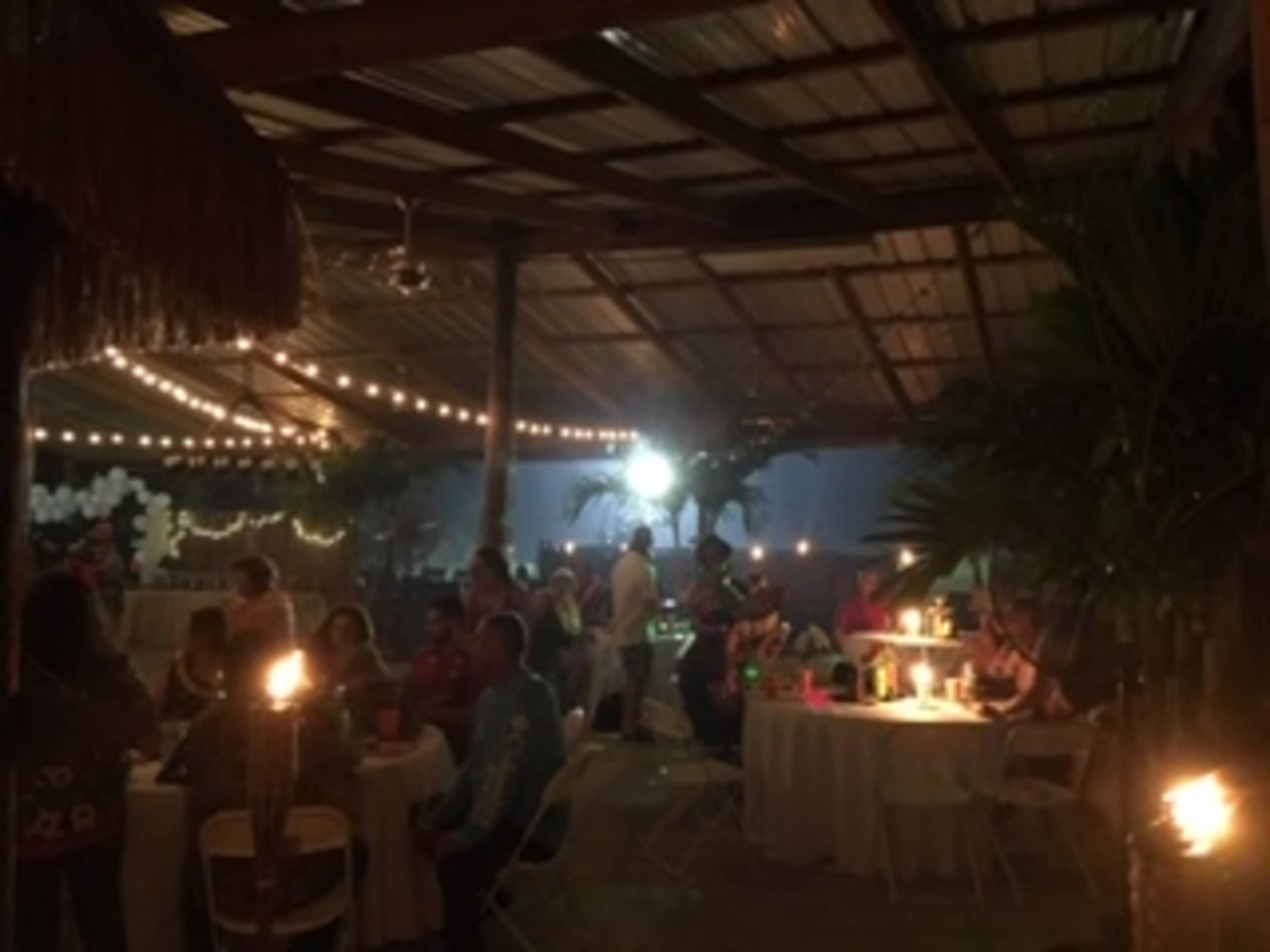 The image shows a lively outdoor event space with people gathered around tables, illuminated by string lights and tiki torches.