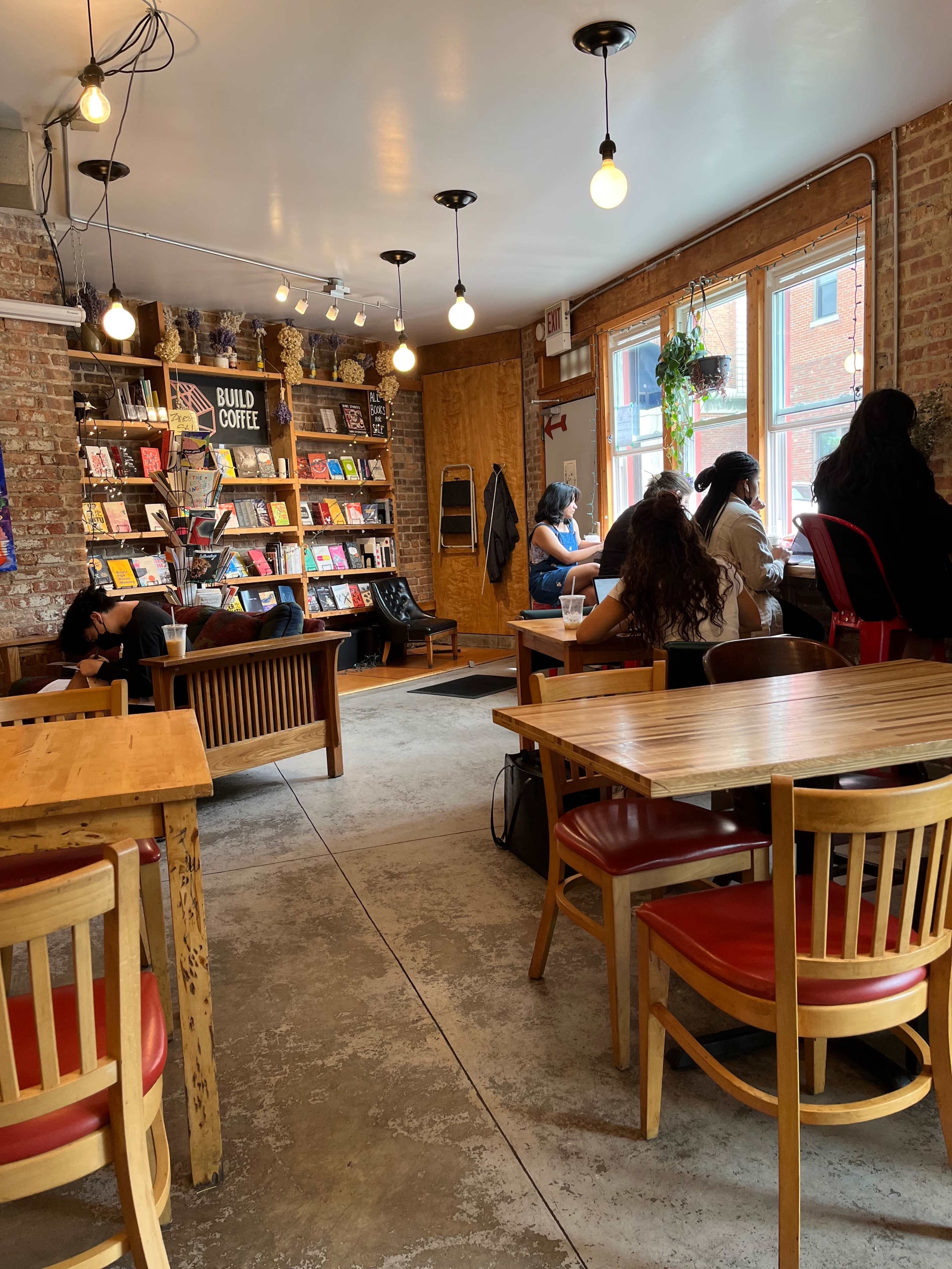A cozy café features wooden tables and chairs, bookshelves filled with books, and customers seated at tables and near windows.