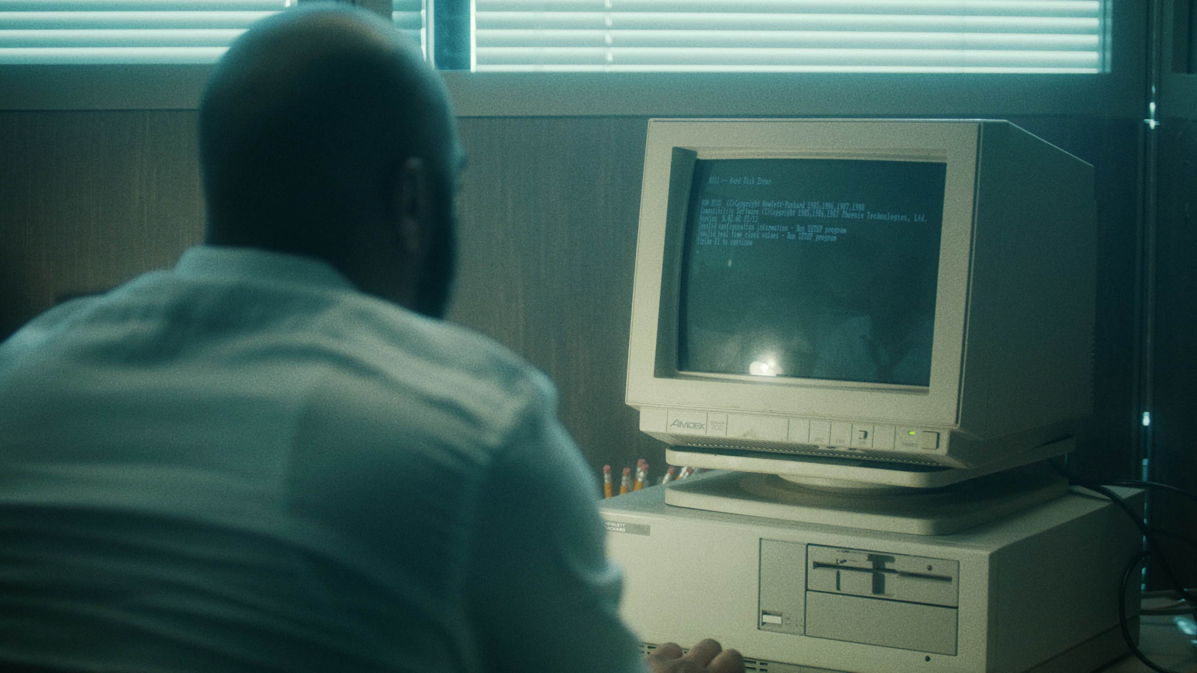 A person sits at a desk, facing an old computer displaying green text on a black screen in a dimly lit room.