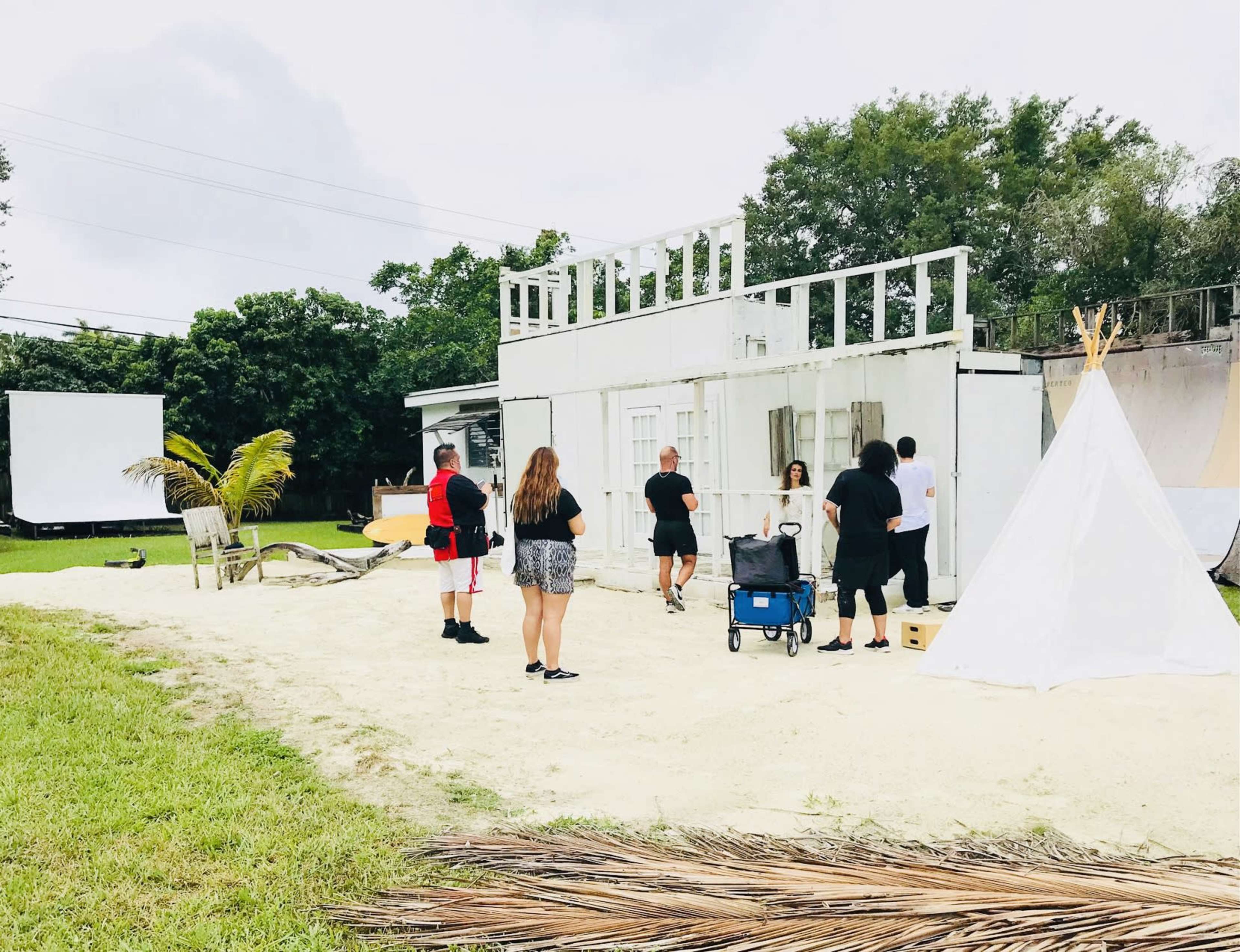 A group of people is preparing a photoshoot in an outdoor area featuring a partially constructed building, a teepee, and various props on the sand.