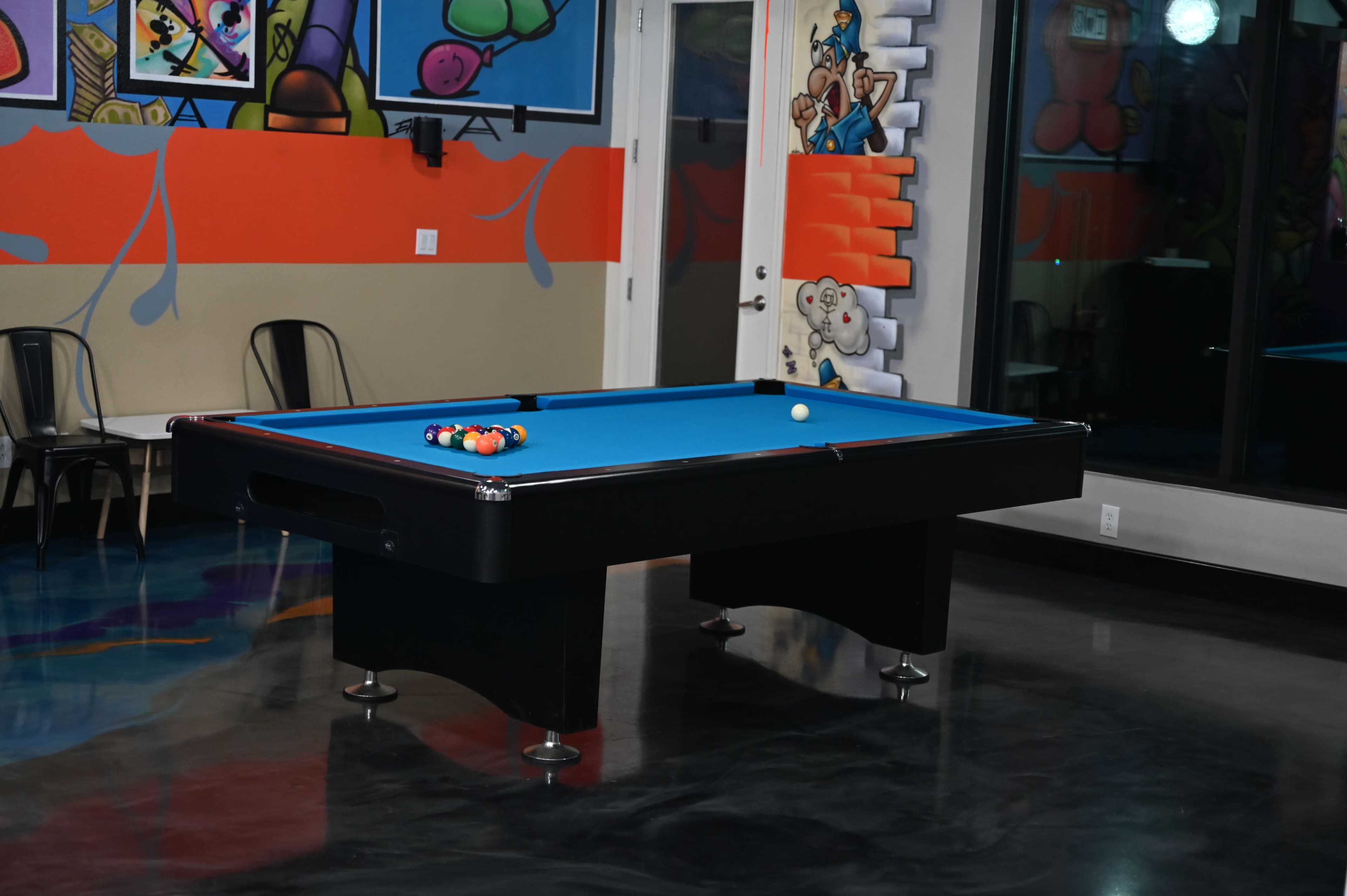 A blue pool table with a cue ball and a set of colored billiard balls is positioned in a brightly decorated room featuring colorful murals.