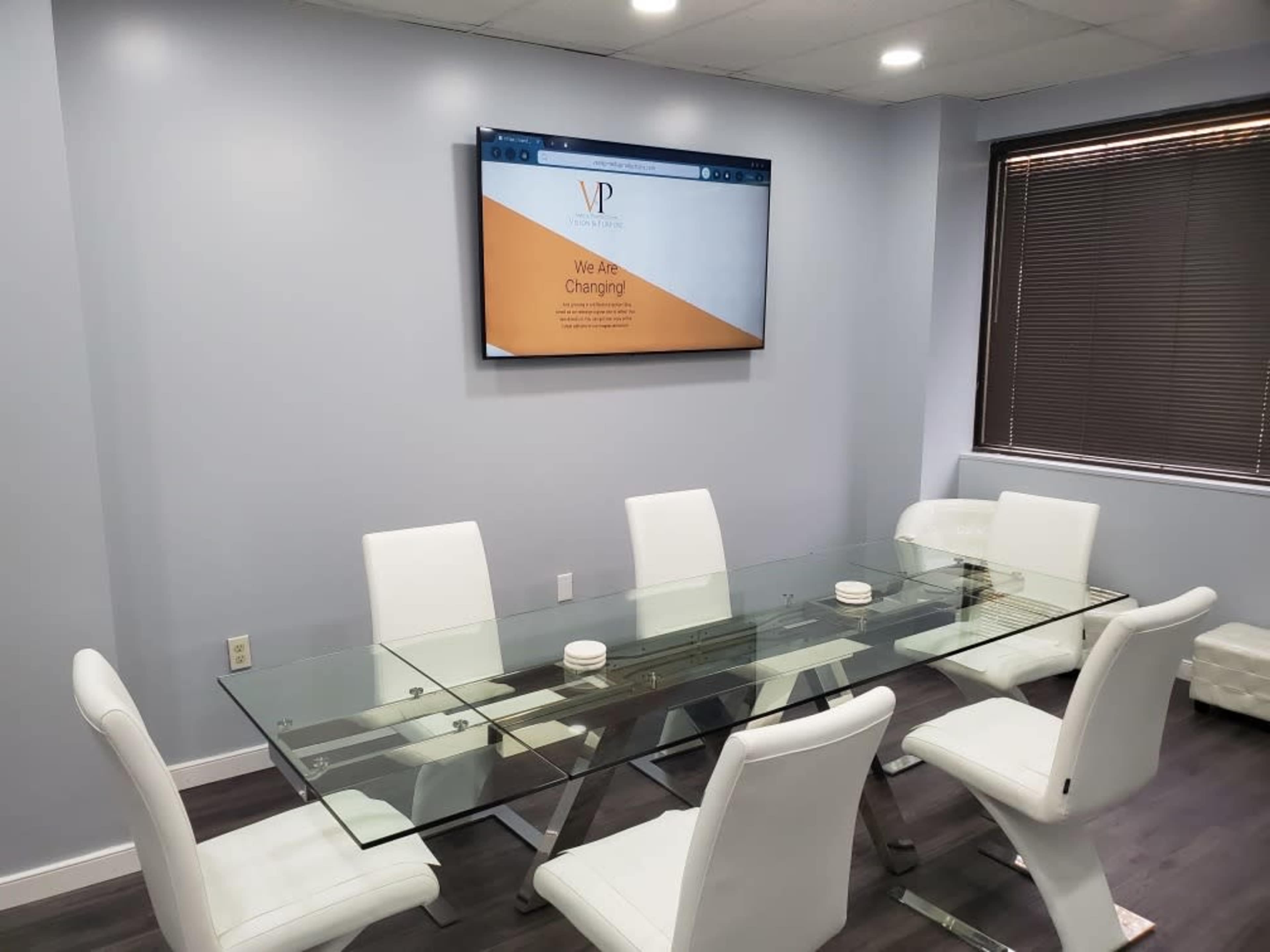 A modern conference room features a glass table surrounded by white chairs, with a large screen displaying a message on the wall.