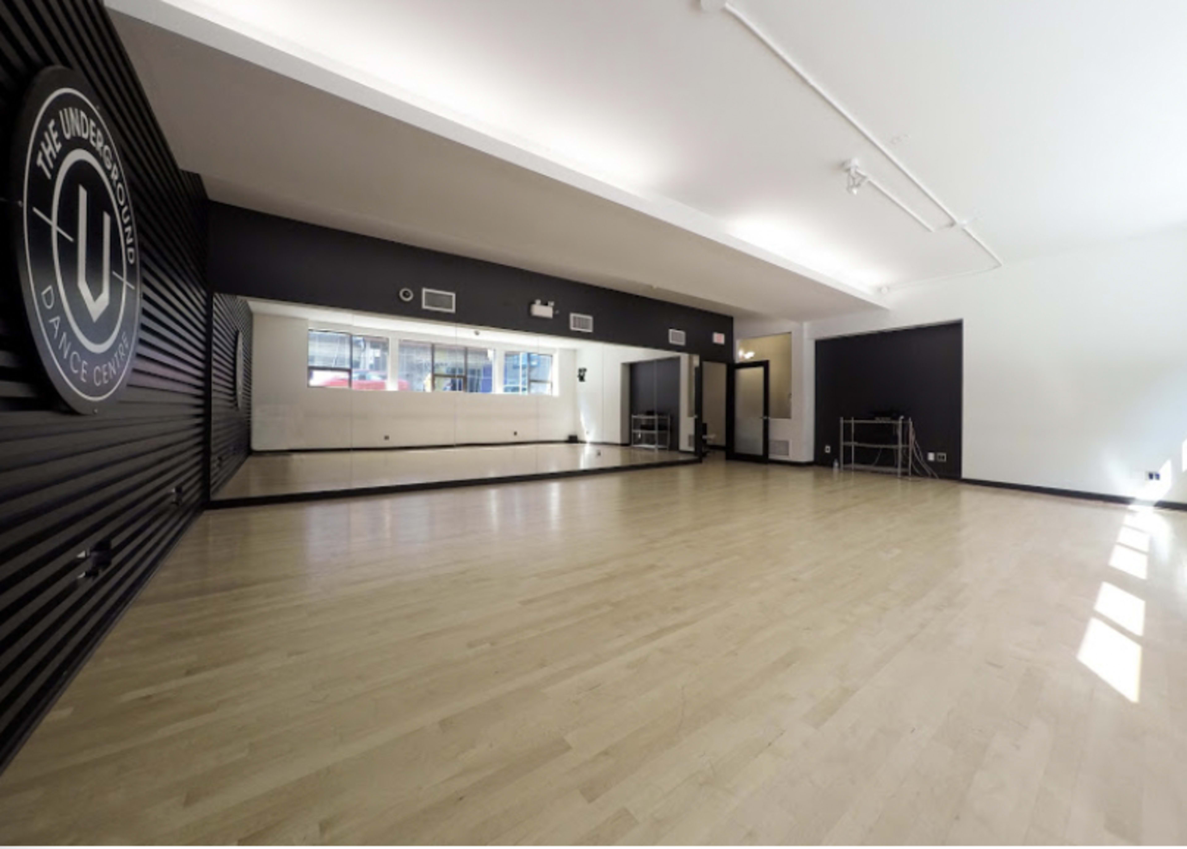 A spacious dance studio features a large mirror along one wall and wooden flooring, with a logo displayed on the opposite wall.
