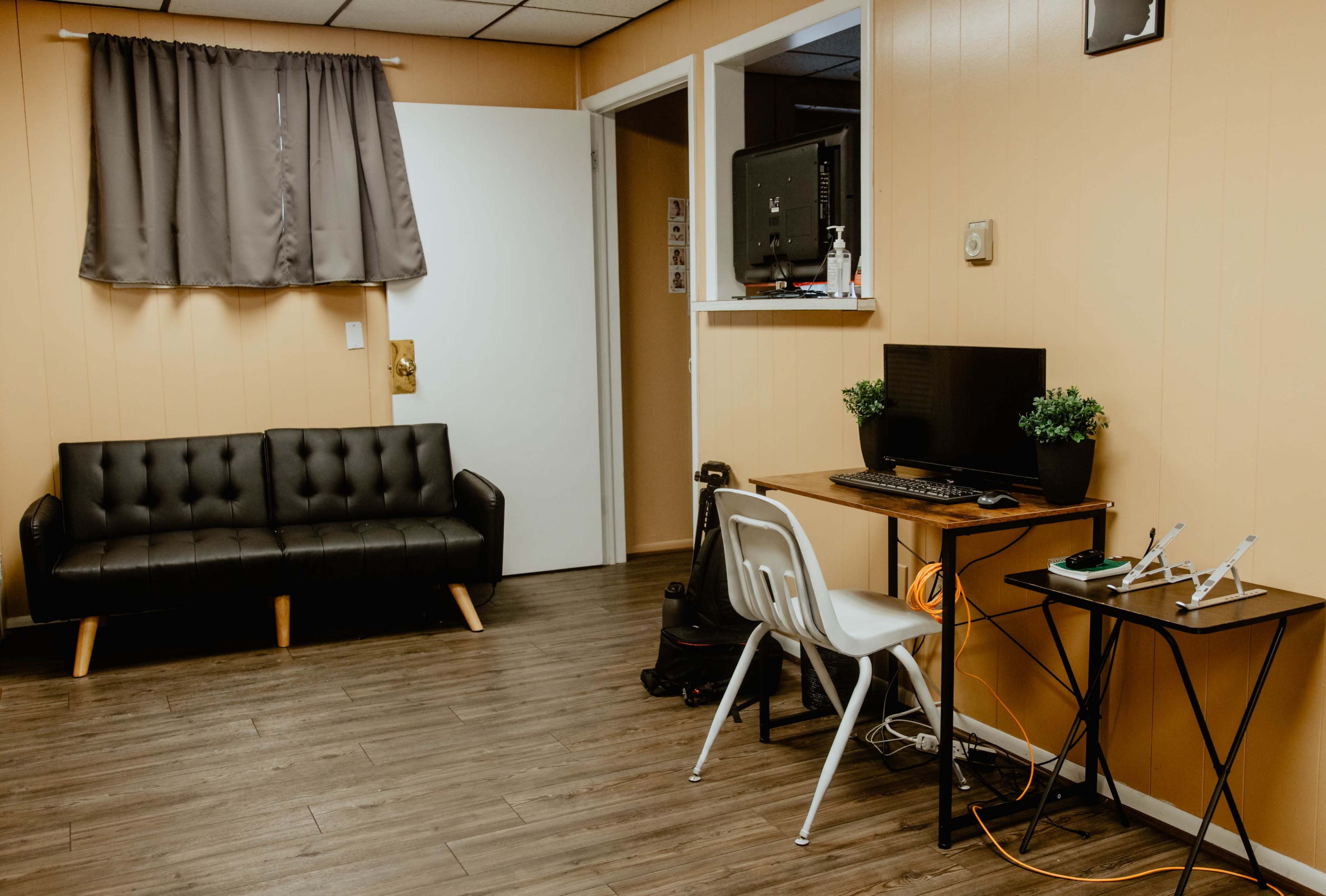 A small room with a black couch, a desk with a computer setup, a white chair, and a door leading to another area, all featuring light brown walls and wooden flooring.