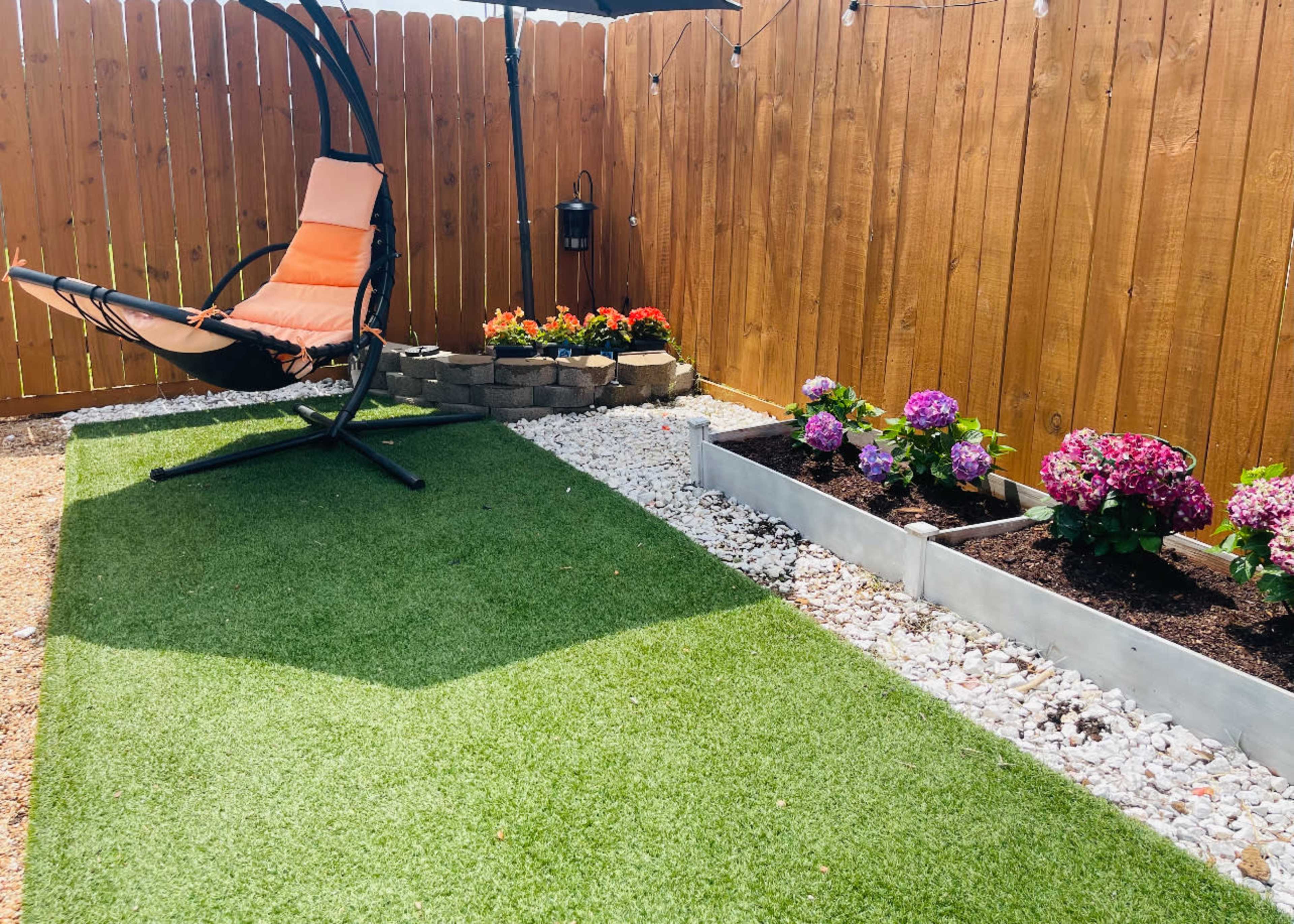 The image shows a small outdoor space featuring a hanging chair, flower beds with blooming plants, and artificial grass next to a wooden fence.