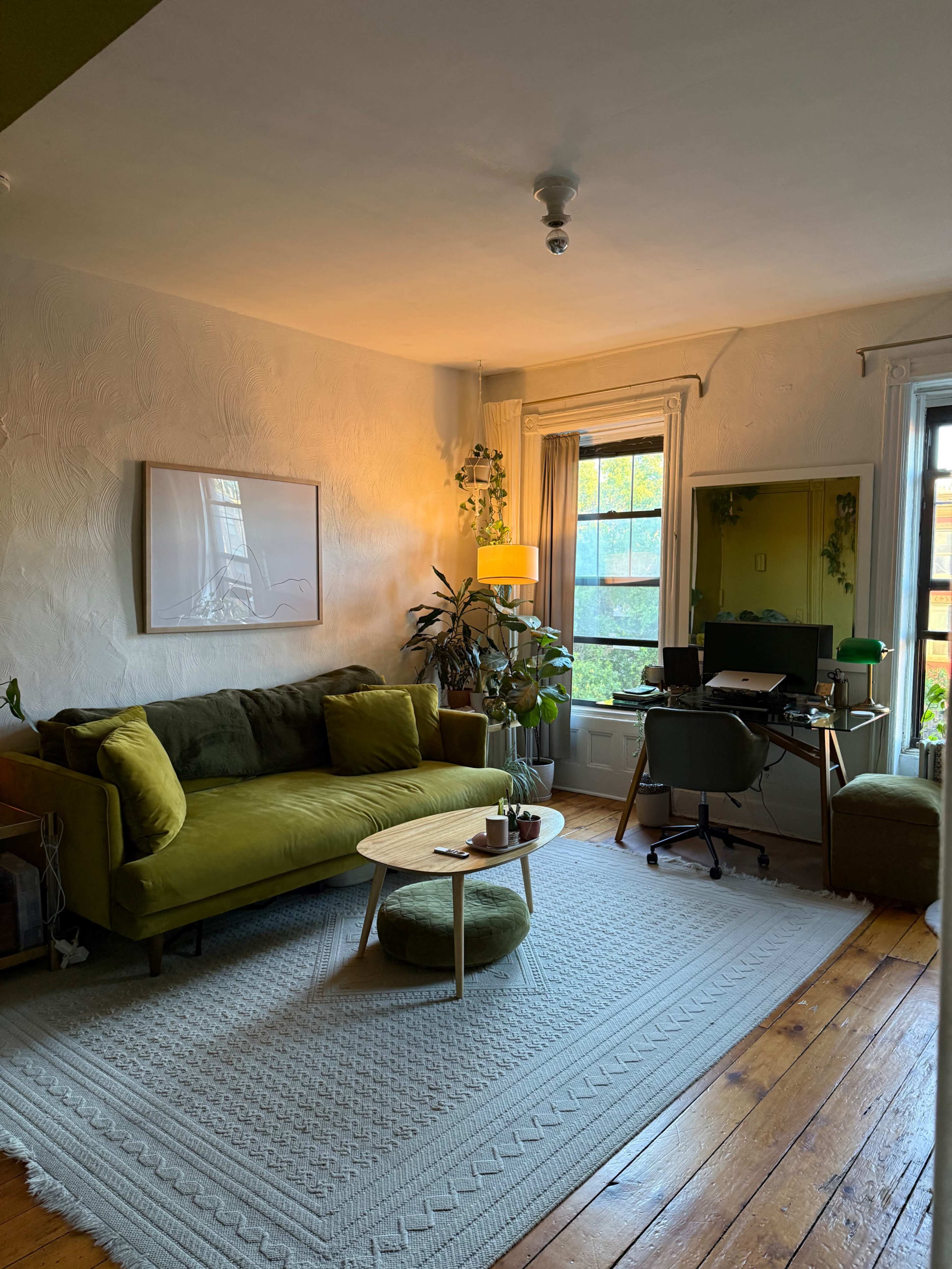 The image shows a cozy living room featuring a green sofa, a round coffee table with several plants, and a desk in the corner by a large window.