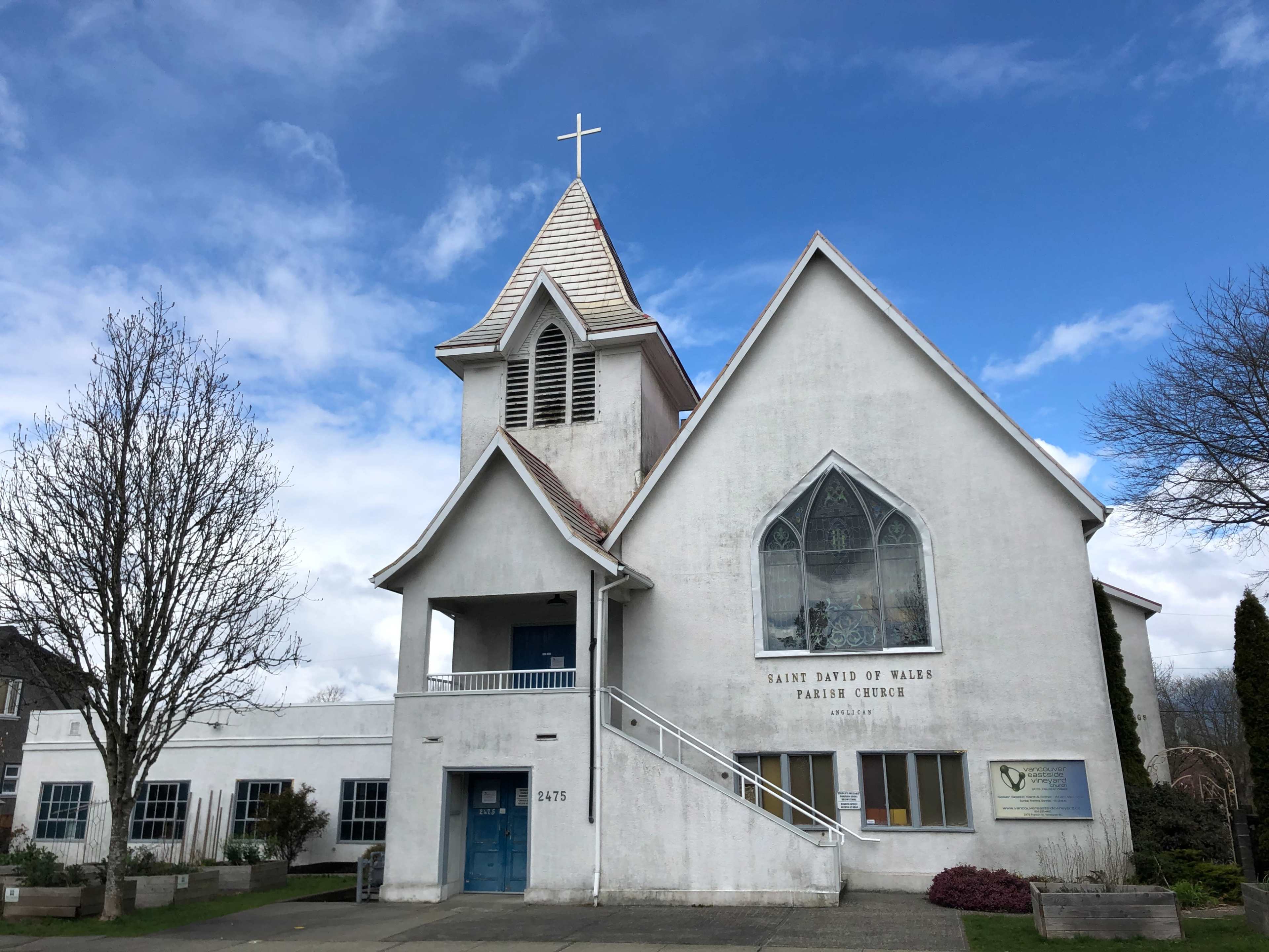 A white church building with a tall steeple, large windows, and a cross on top against a partly cloudy sky.