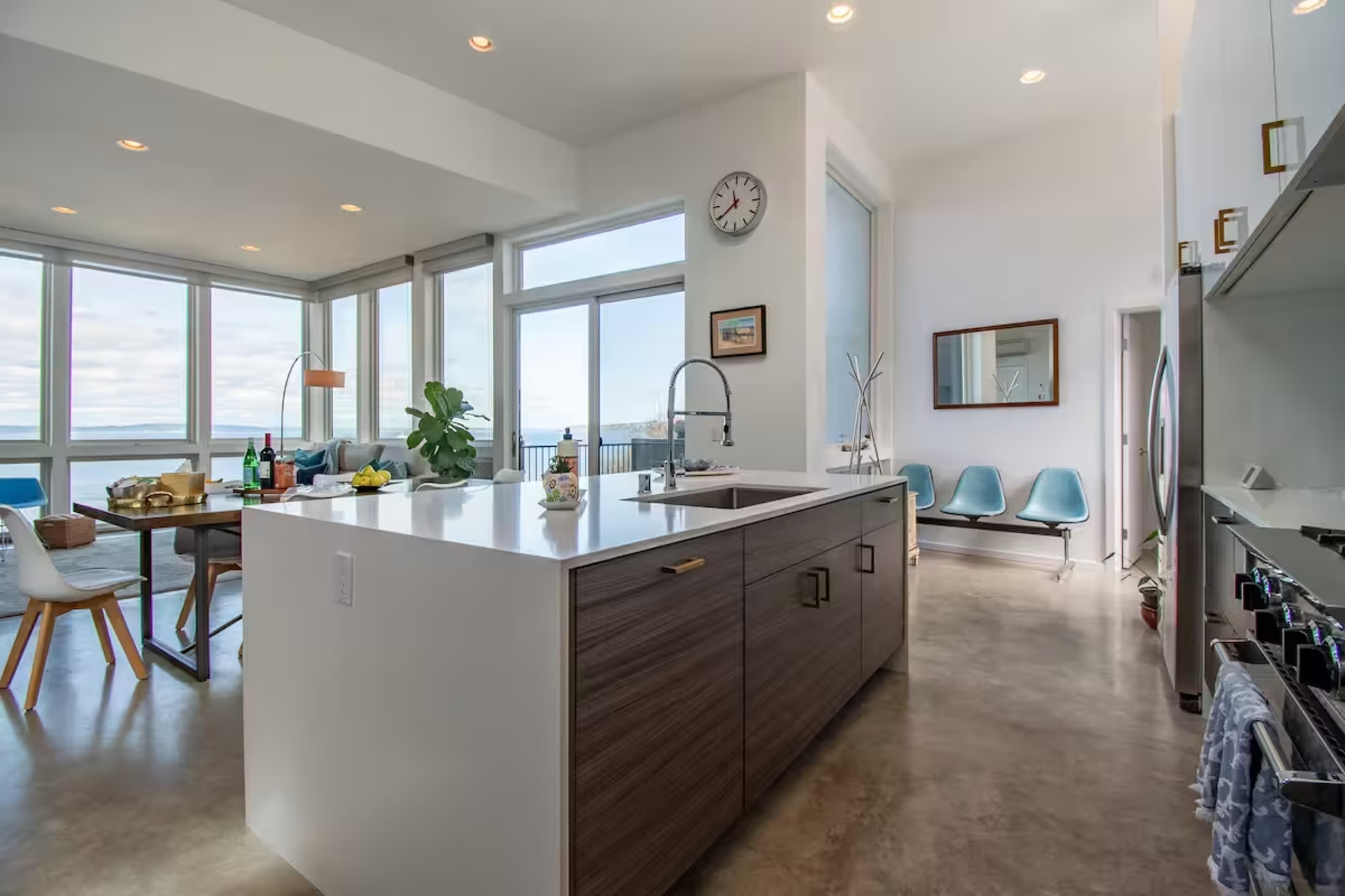 The image shows a modern kitchen with an island, featuring large windows that provide views of the outdoors, and a dining area with a wooden table and chairs.