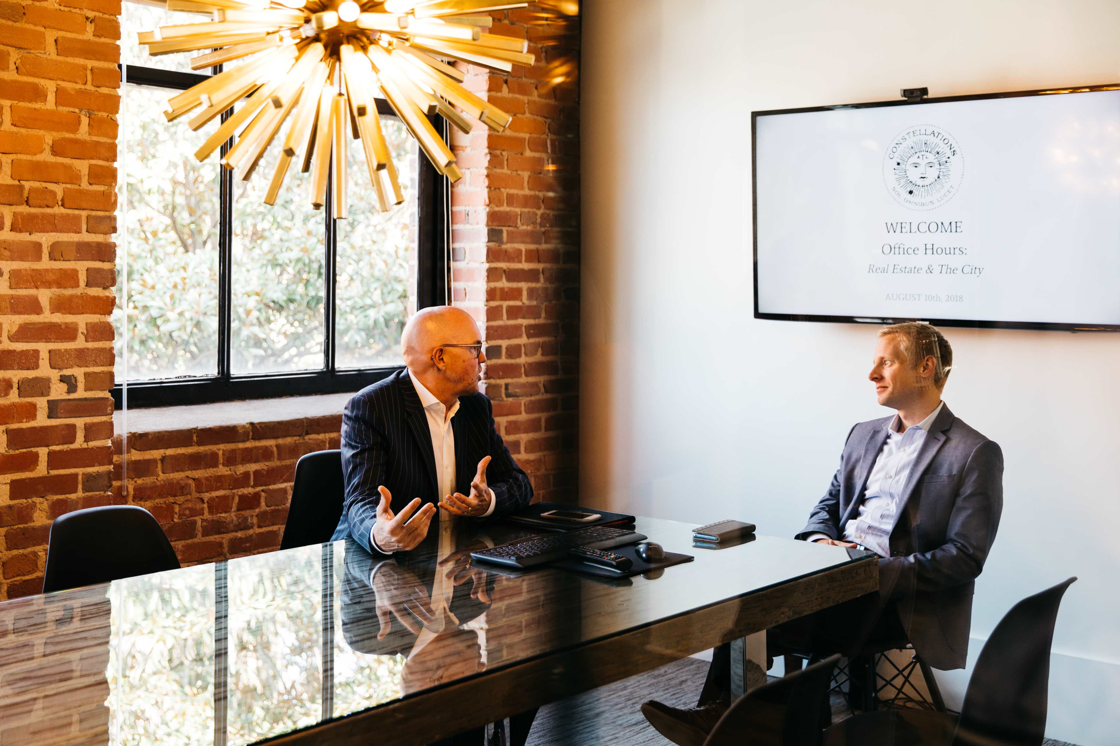 Two men are engaged in conversation at a glass-topped table in a modern office with exposed brick walls and a large light fixture.