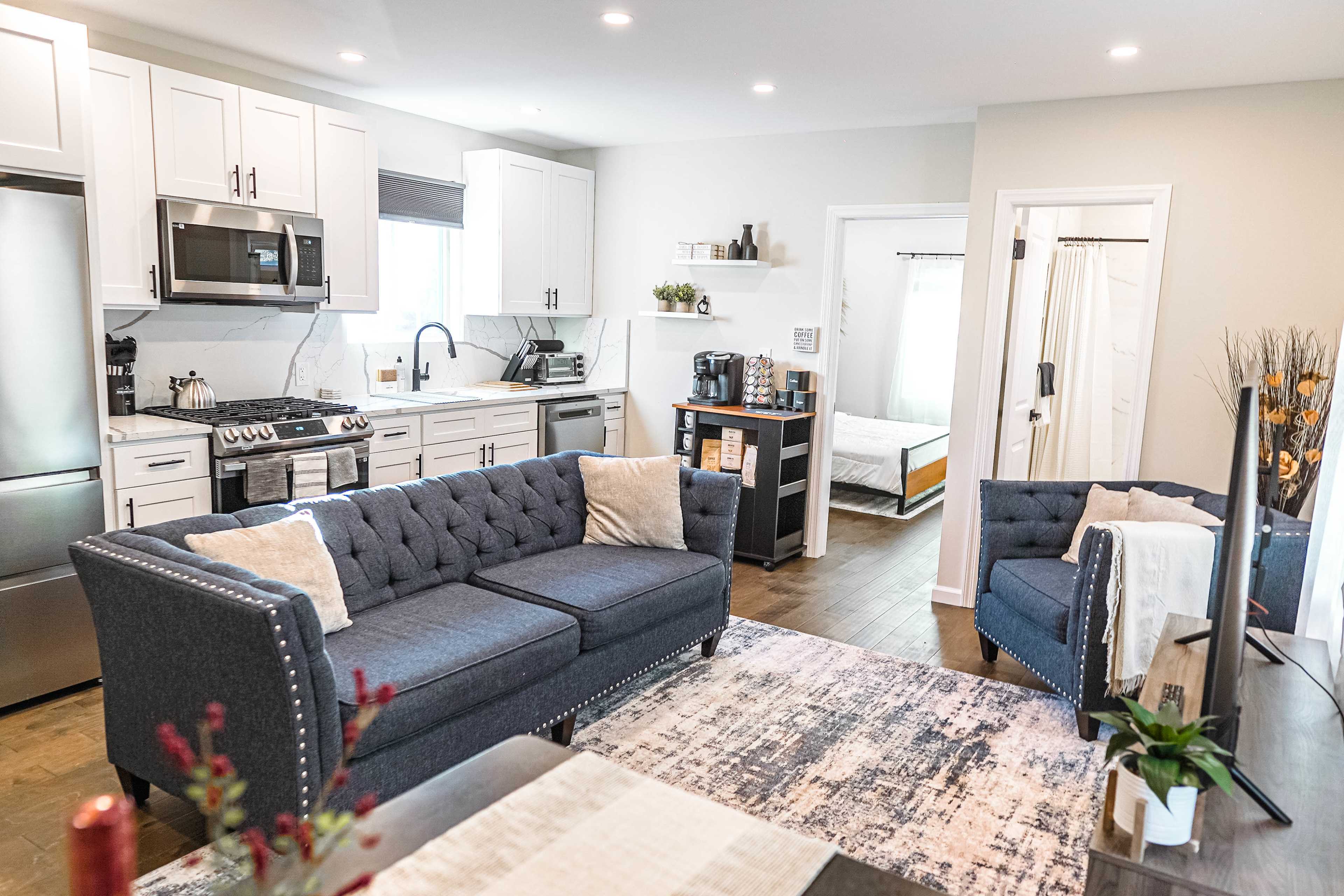 A modern living room features a blue sectional sofa, a pair of armchairs, and a kitchen area with stainless steel appliances and white cabinetry.