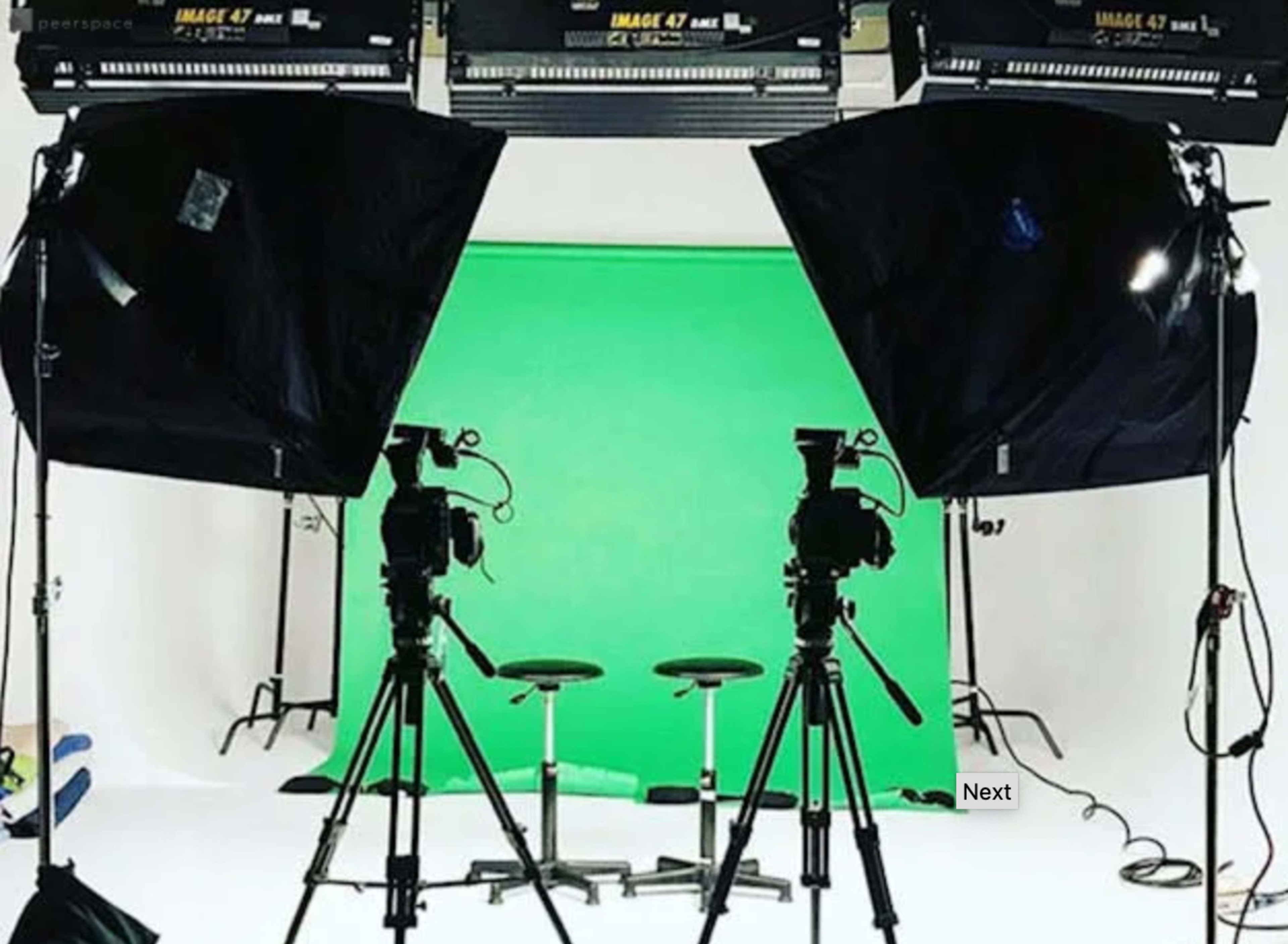 A studio setup with two cameras on tripods facing a green backdrop and two stools placed in front.