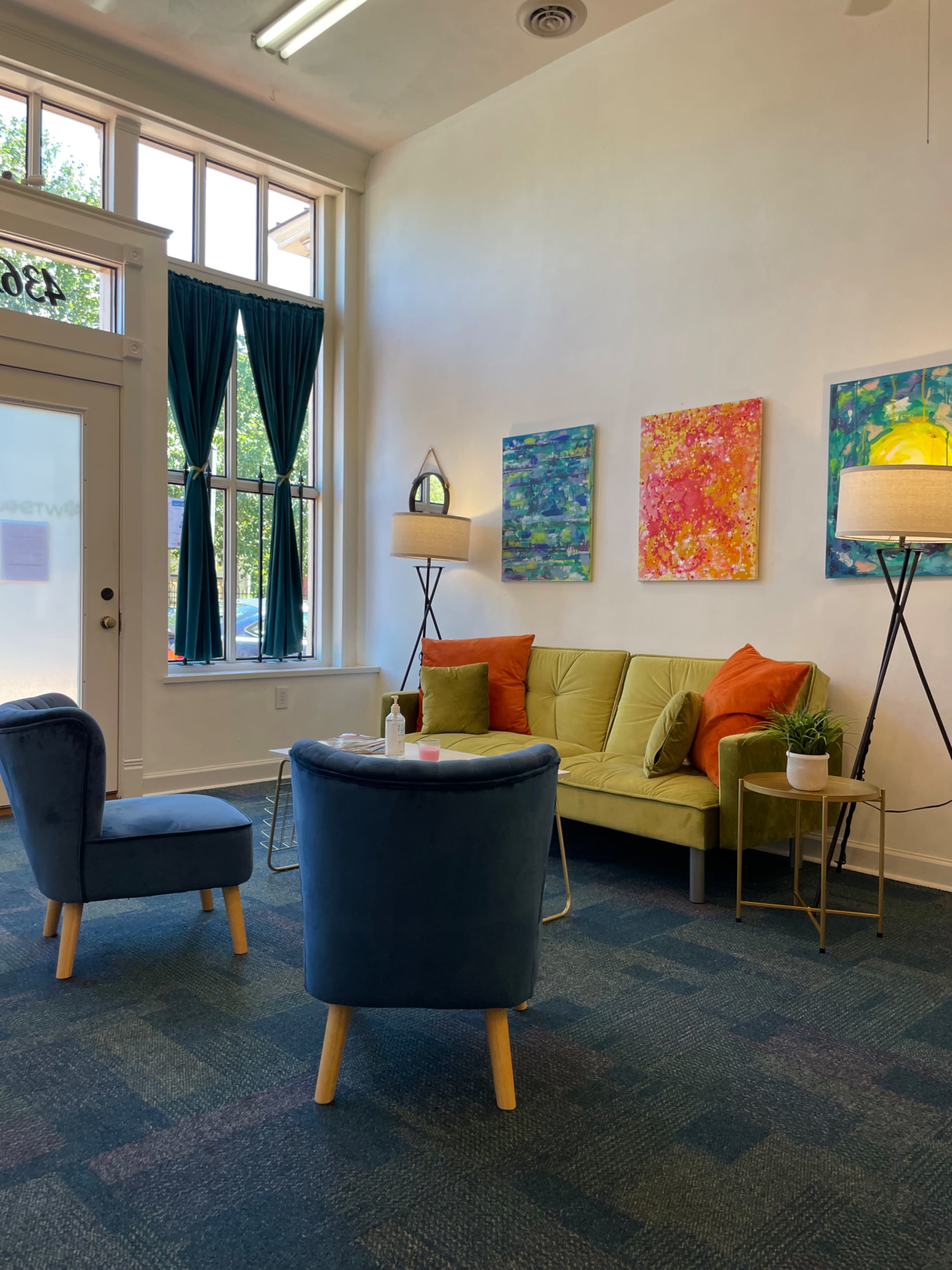 A waiting area with a green sofa, colorful wall art, and two lamps, accompanied by a blue armchair and a coffee table.