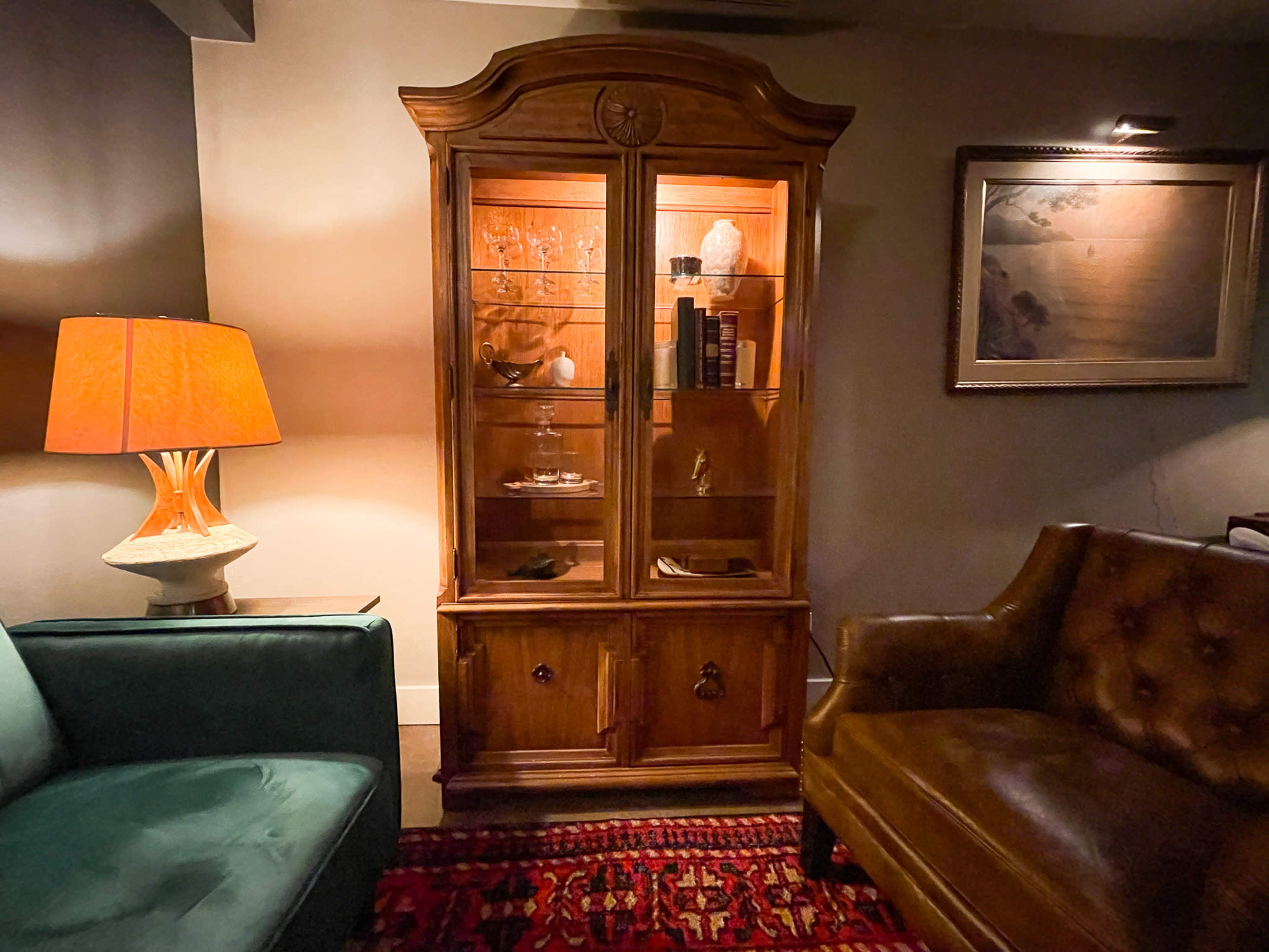 The image features a wooden display cabinet illuminated inside, positioned between a green sofa and a brown leather chair, with a lamp casting light on one side and a framed picture on the wall in the background.