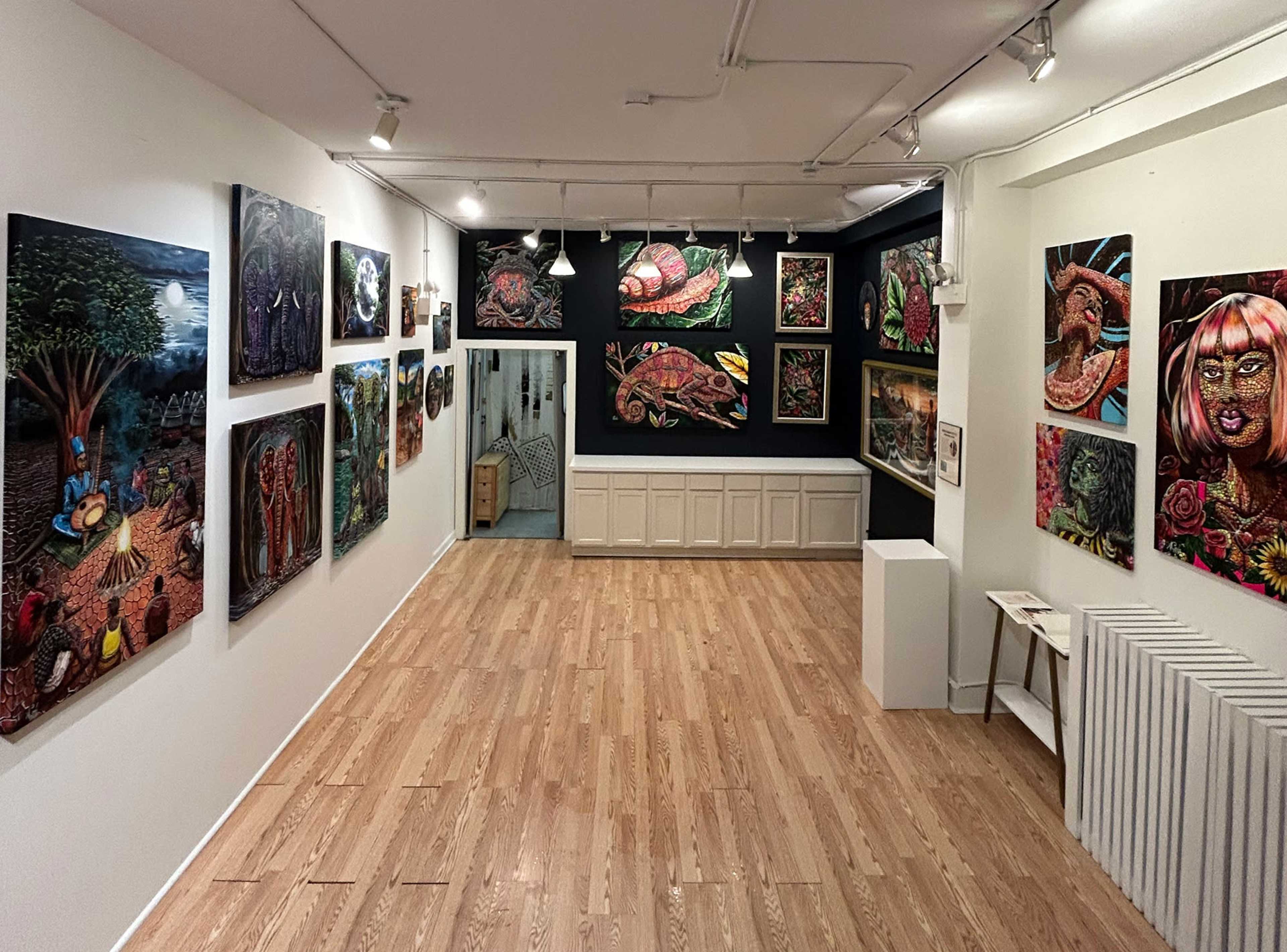 The image shows an art gallery with various colorful paintings displayed on the walls, featuring diverse subjects and styles, with a wooden floor and bright lighting illuminating the space.