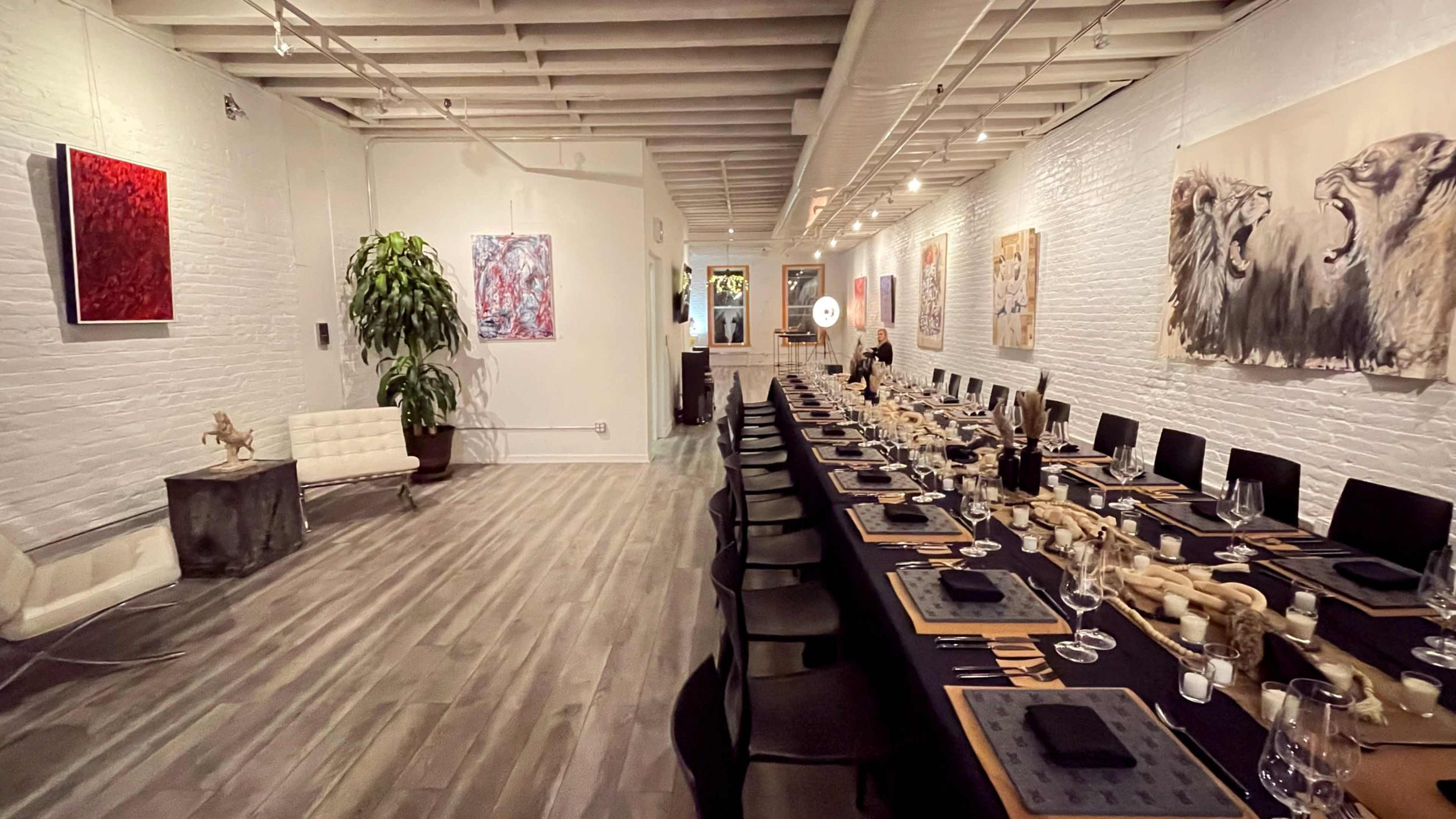 The image shows a long dining table set for a meal in a spacious, well-lit gallery with various artworks displayed on the walls.