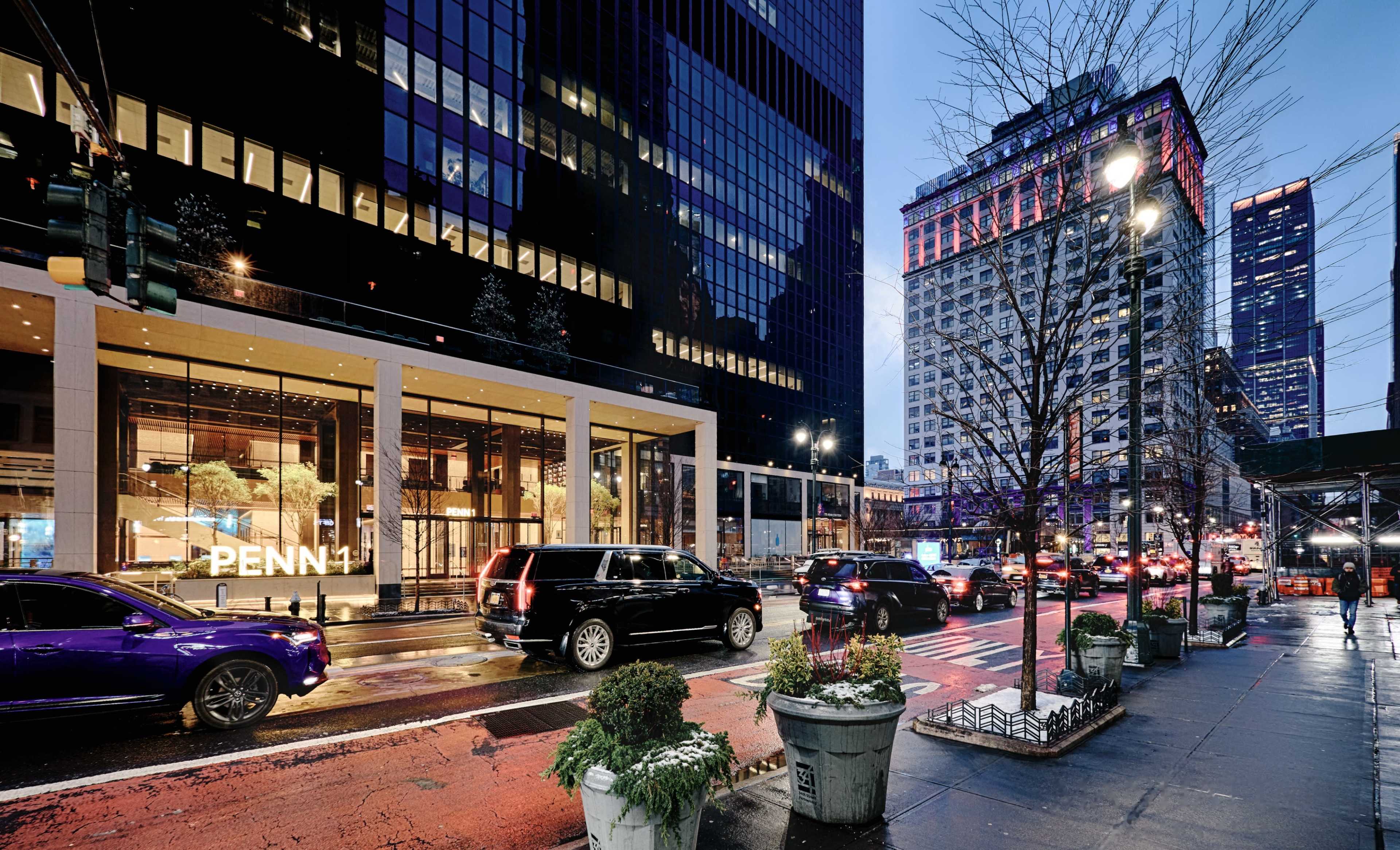 The image shows a busy city street with traffic, modern buildings, and planters along the sidewalk in a twilight setting.