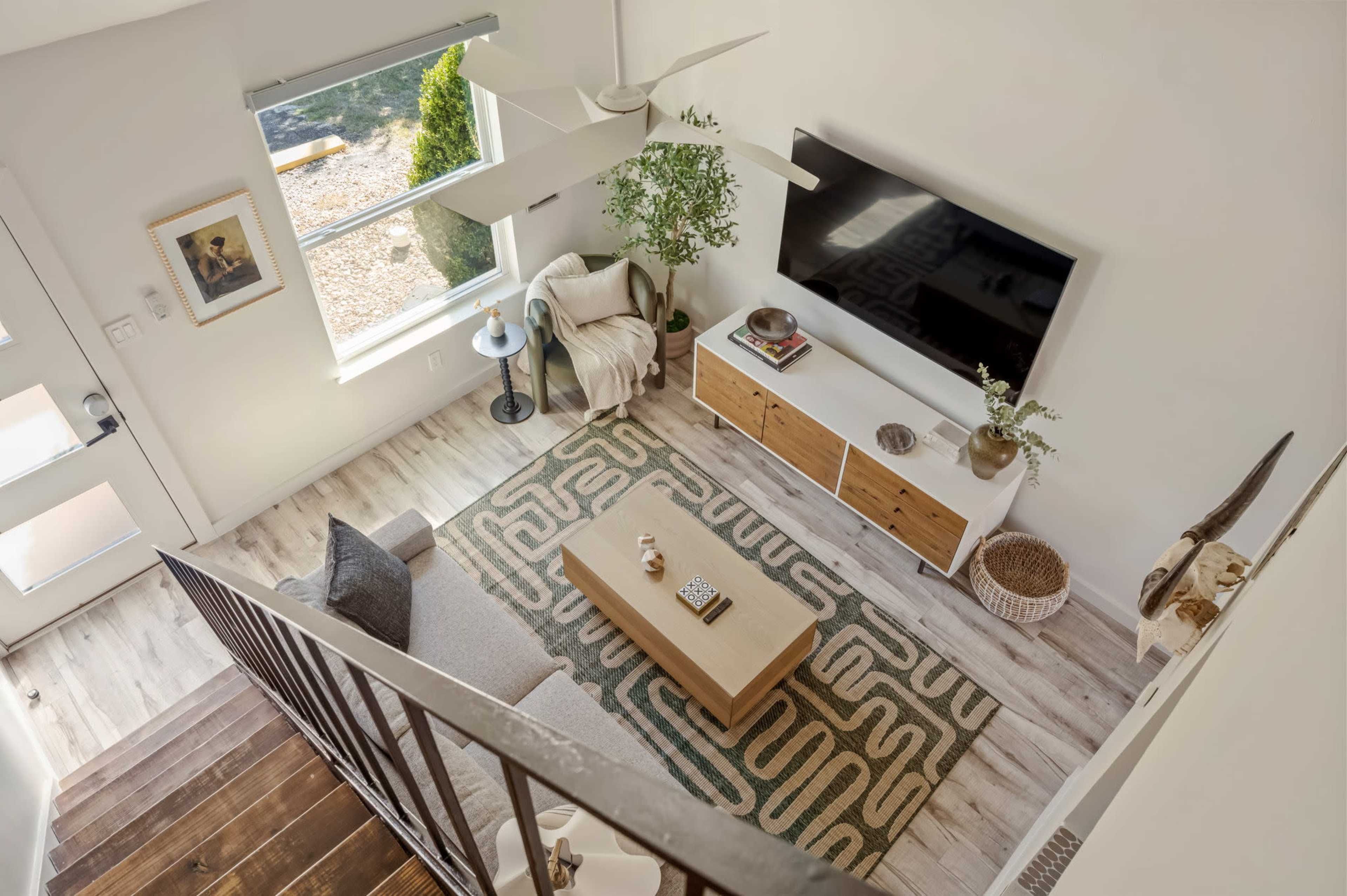 A modern living room features a light-colored sofa, wooden coffee table, and a large wall-mounted television, with a view of an outdoor area through a window.