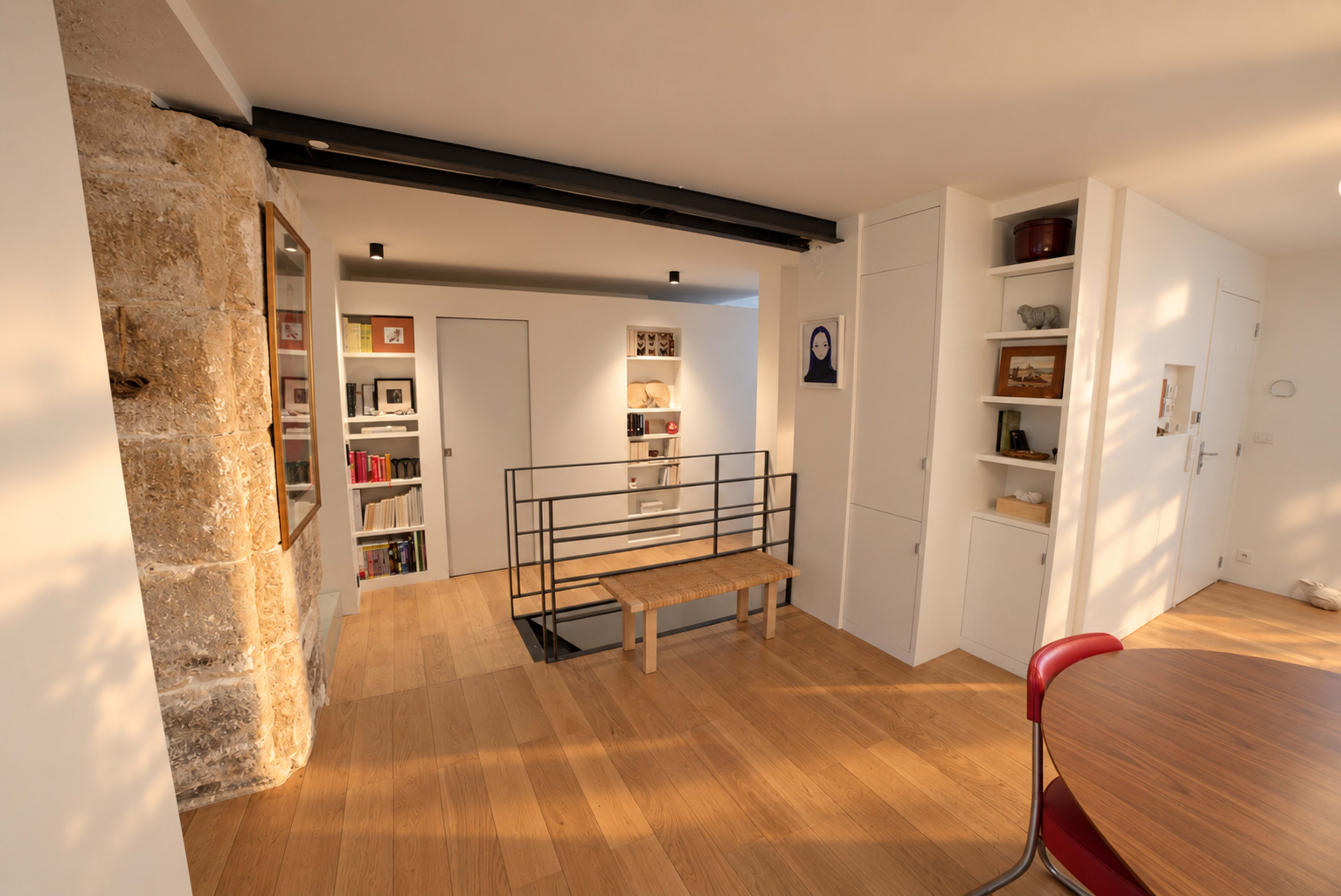 The image shows a modern interior space featuring a wooden floor, a staircase with a black railing, built-in bookshelves, and a circular dining table surrounded by red chairs.