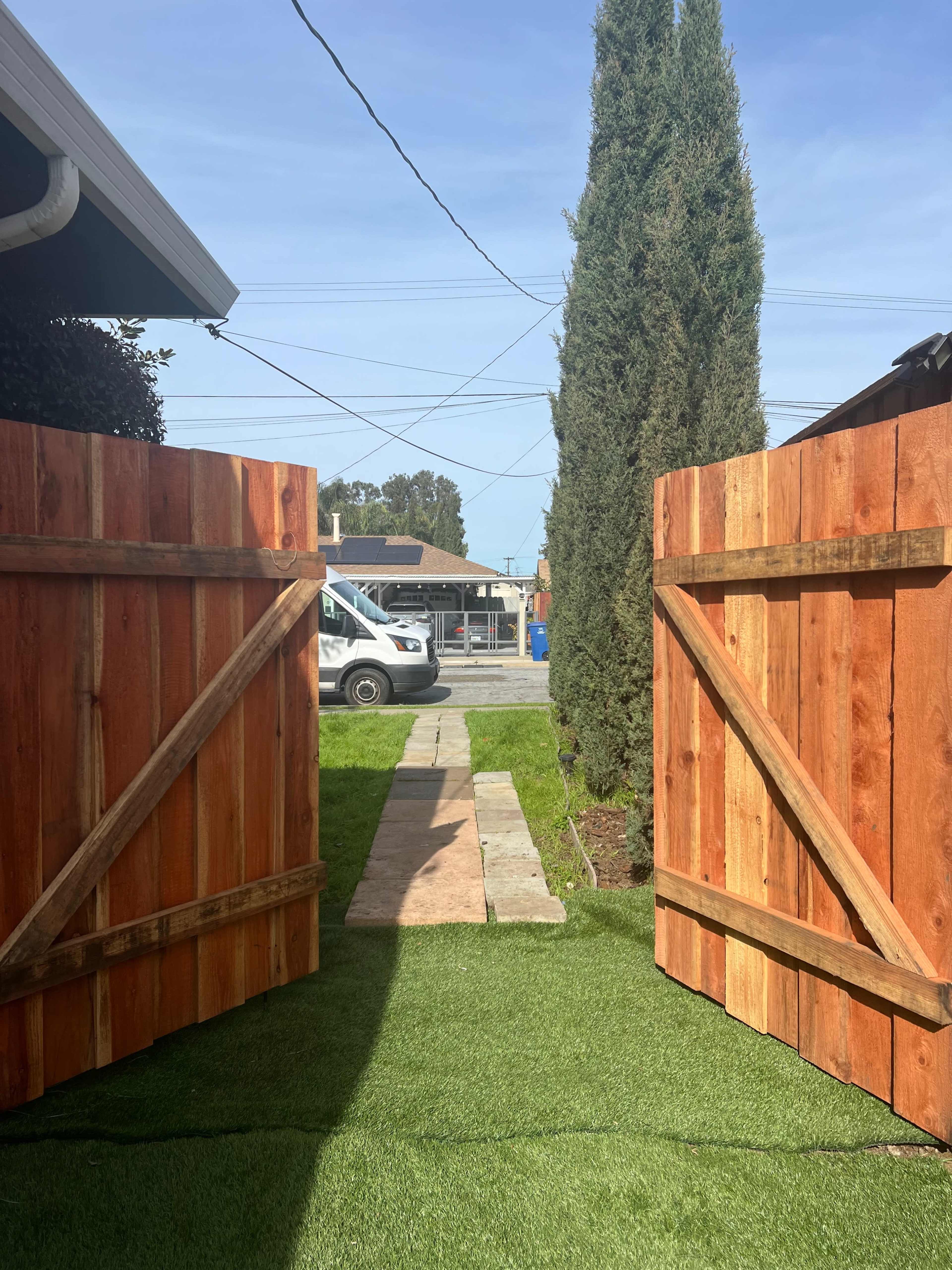 A pair of open wooden gates leading to a pathway lined with grass and a van parked in the background.