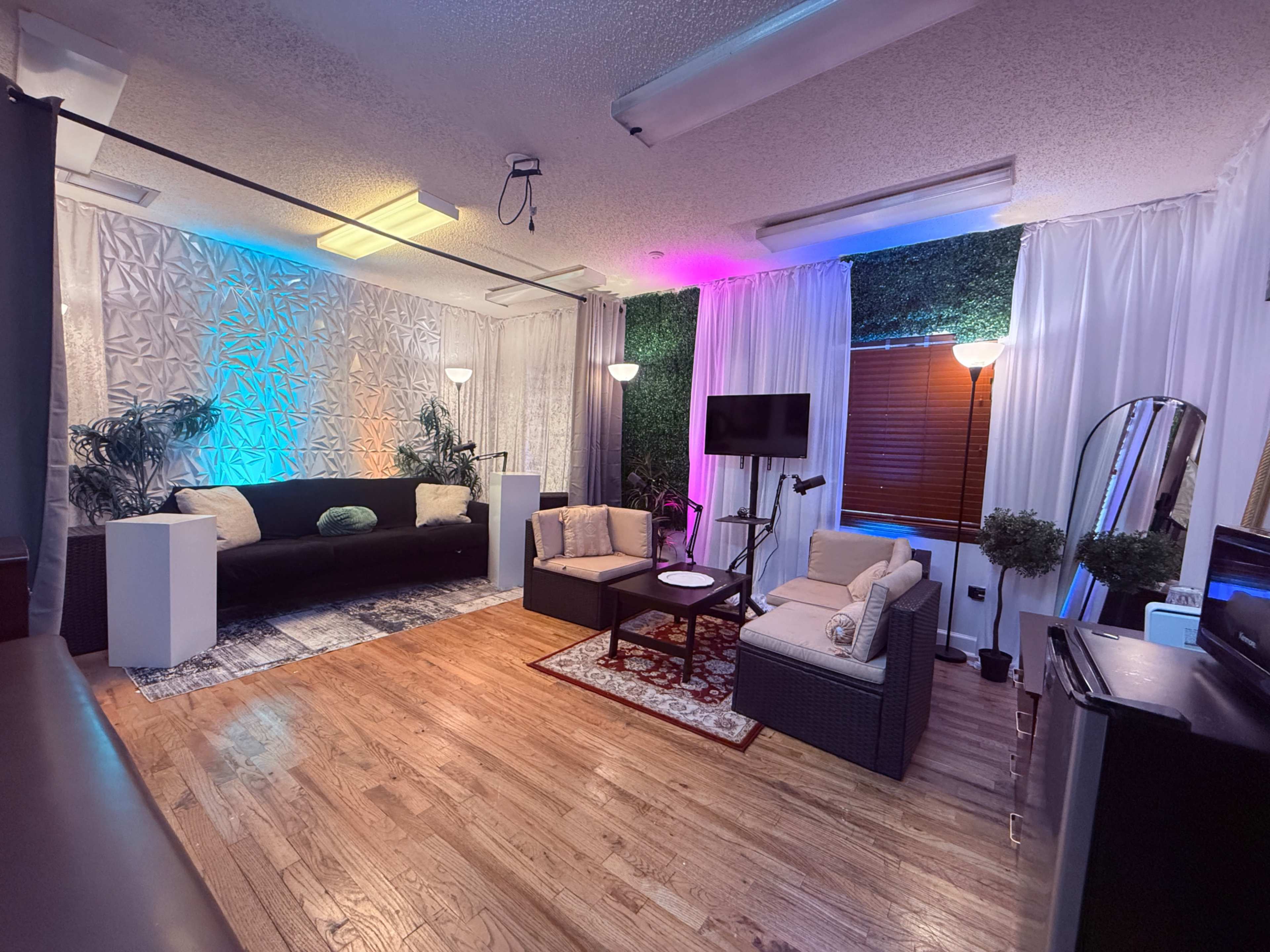 The image shows a modern living space featuring a seating area with a black sofa and two armchairs, accentuated by colorful lighting and decorative plants.