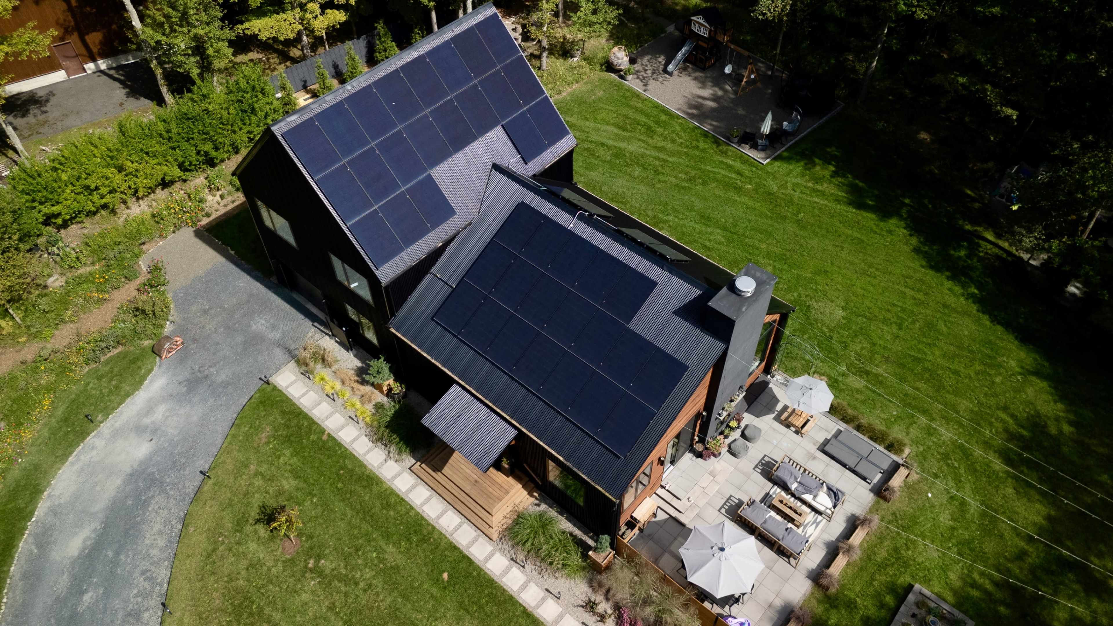 The image shows a modern house with solar panels on the roof, surrounded by a well-maintained lawn and a patio area with outdoor seating.