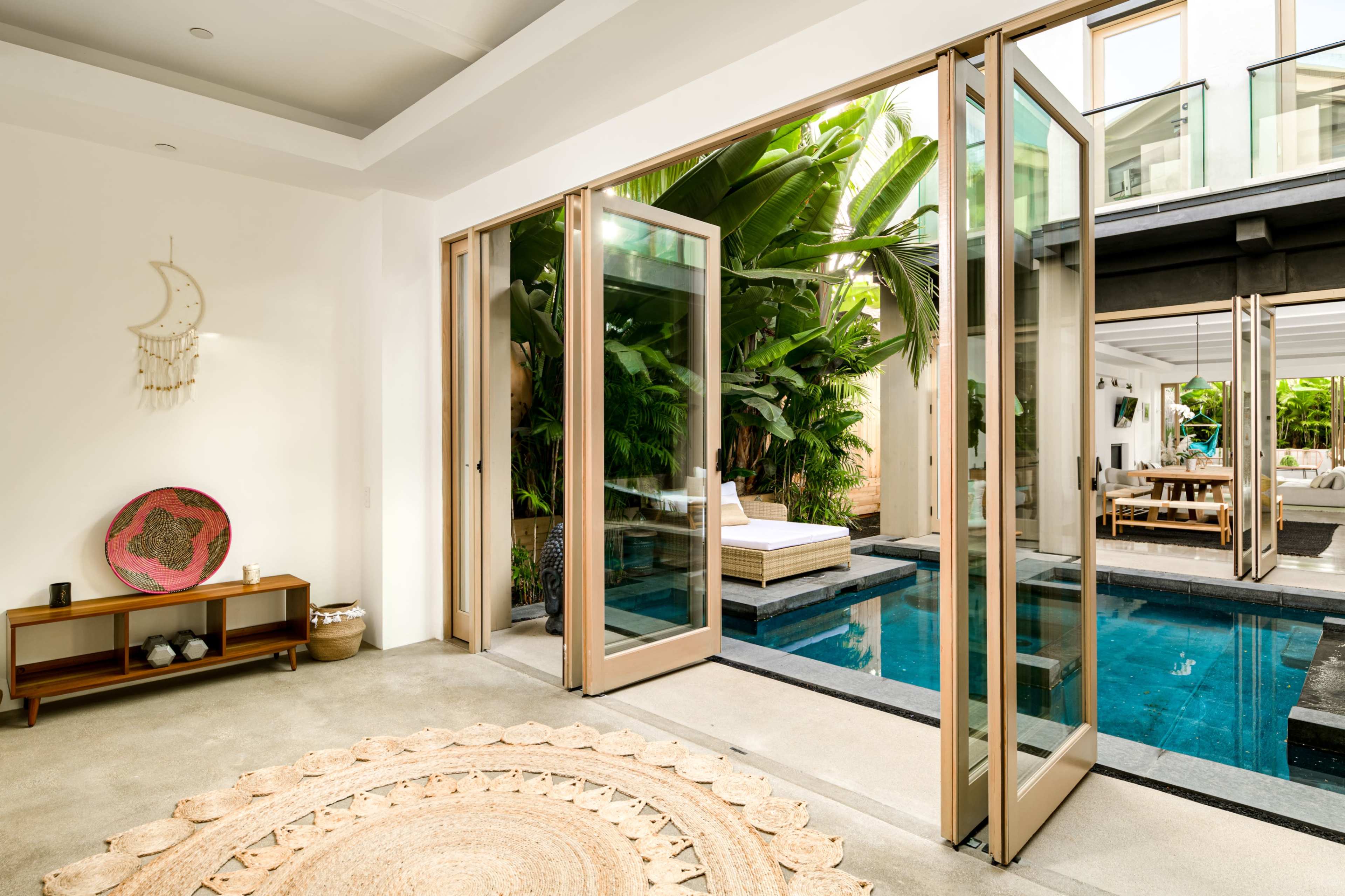 A modern indoor space opens up to a tropical outdoor area featuring a pool and lounge seating surrounded by greenery.