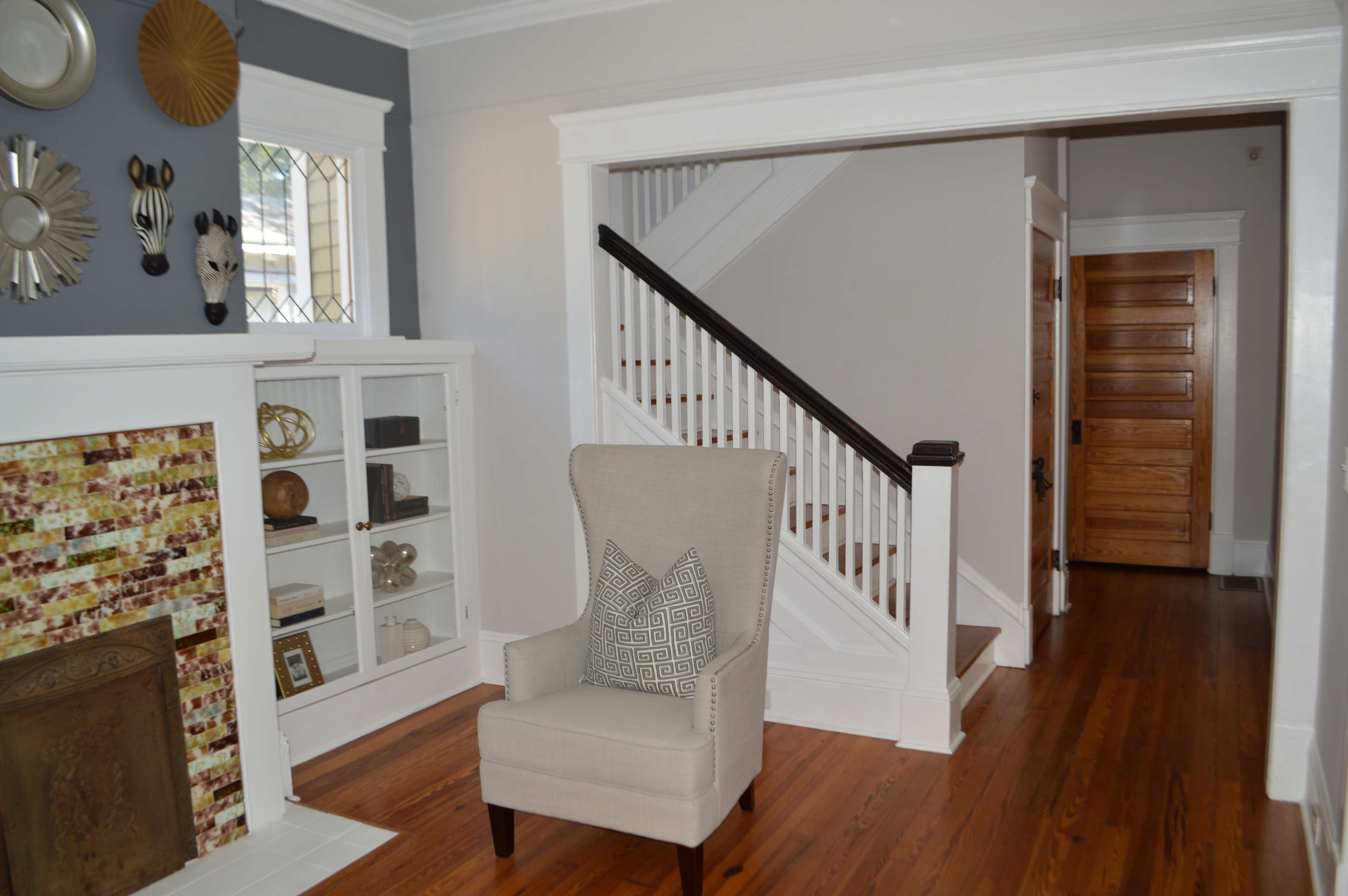 The image shows a living space with a beige armchair in front of a glass-fronted cabinet, a staircase in the background, and wooden floors.