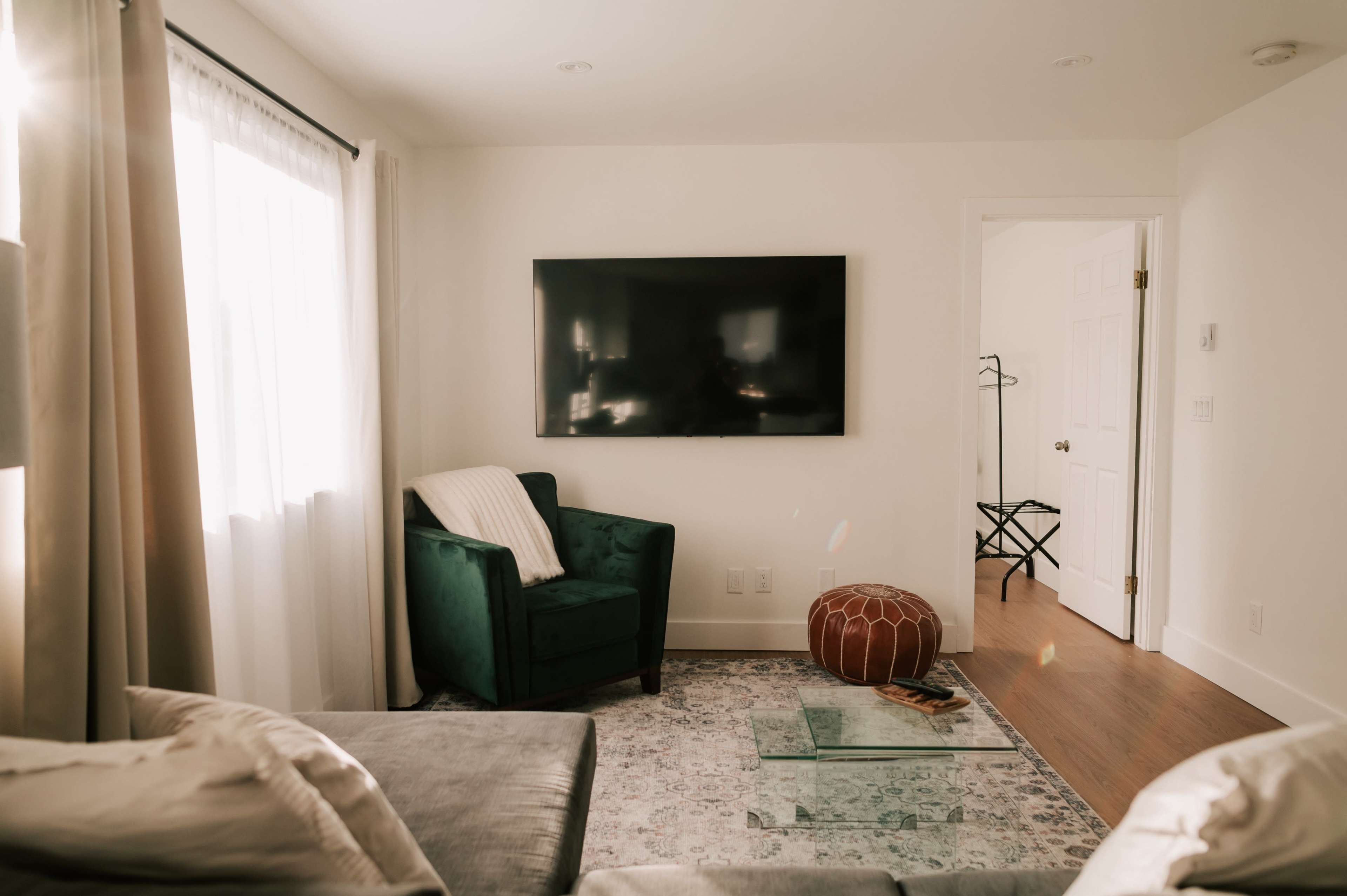 The image shows a modern living room with a green armchair, a large flat-screen TV mounted on the wall, a glass coffee table, and a decorative pouf.