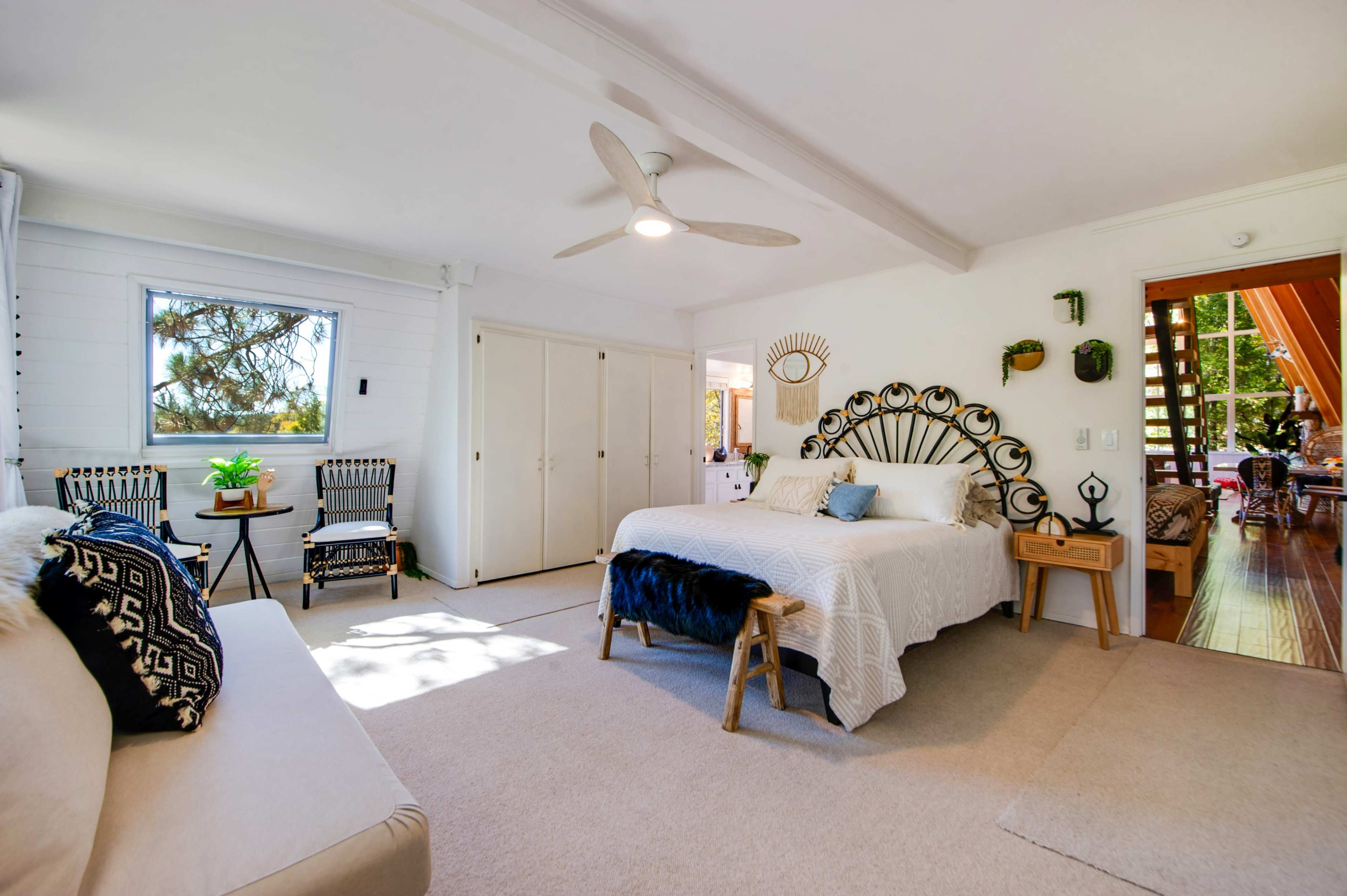 A bright bedroom features a large bed, a seating area, and a doorway leading to an outdoor space, with wood accents and natural light from multiple windows.