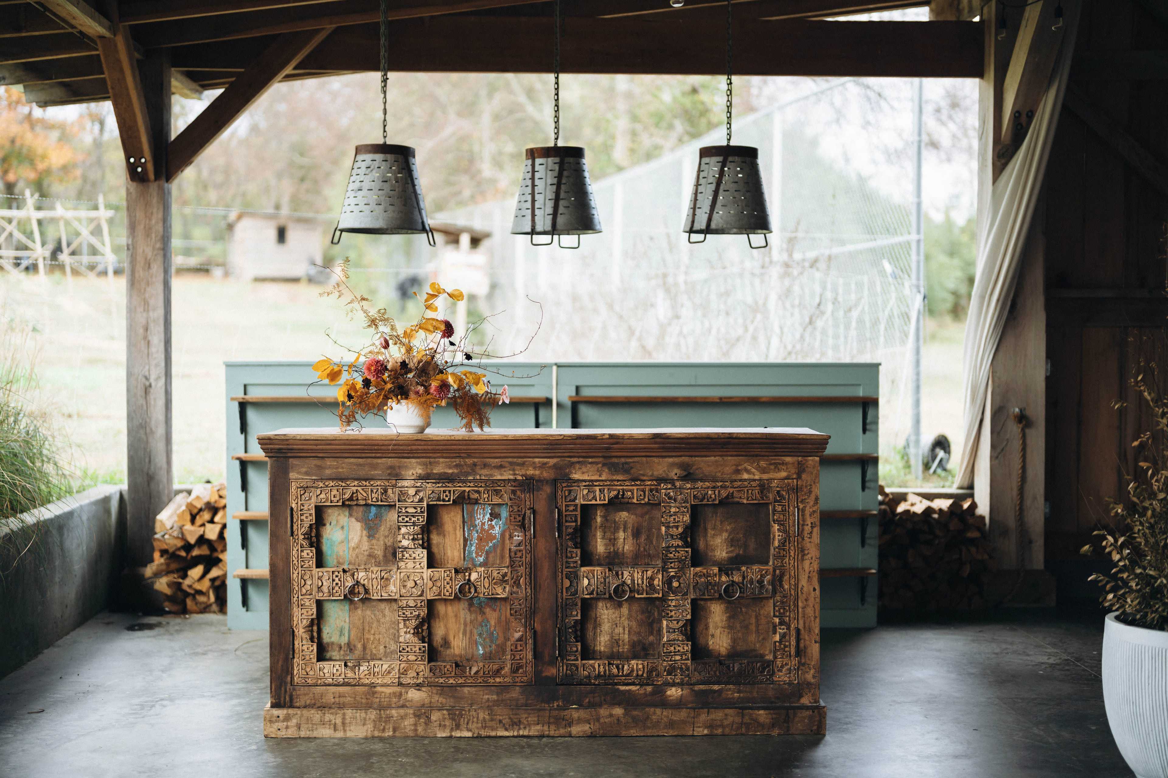 A wooden, intricately carved counter with a floral arrangement on top, set beneath three hanging metal light fixtures in a rustic, open space.