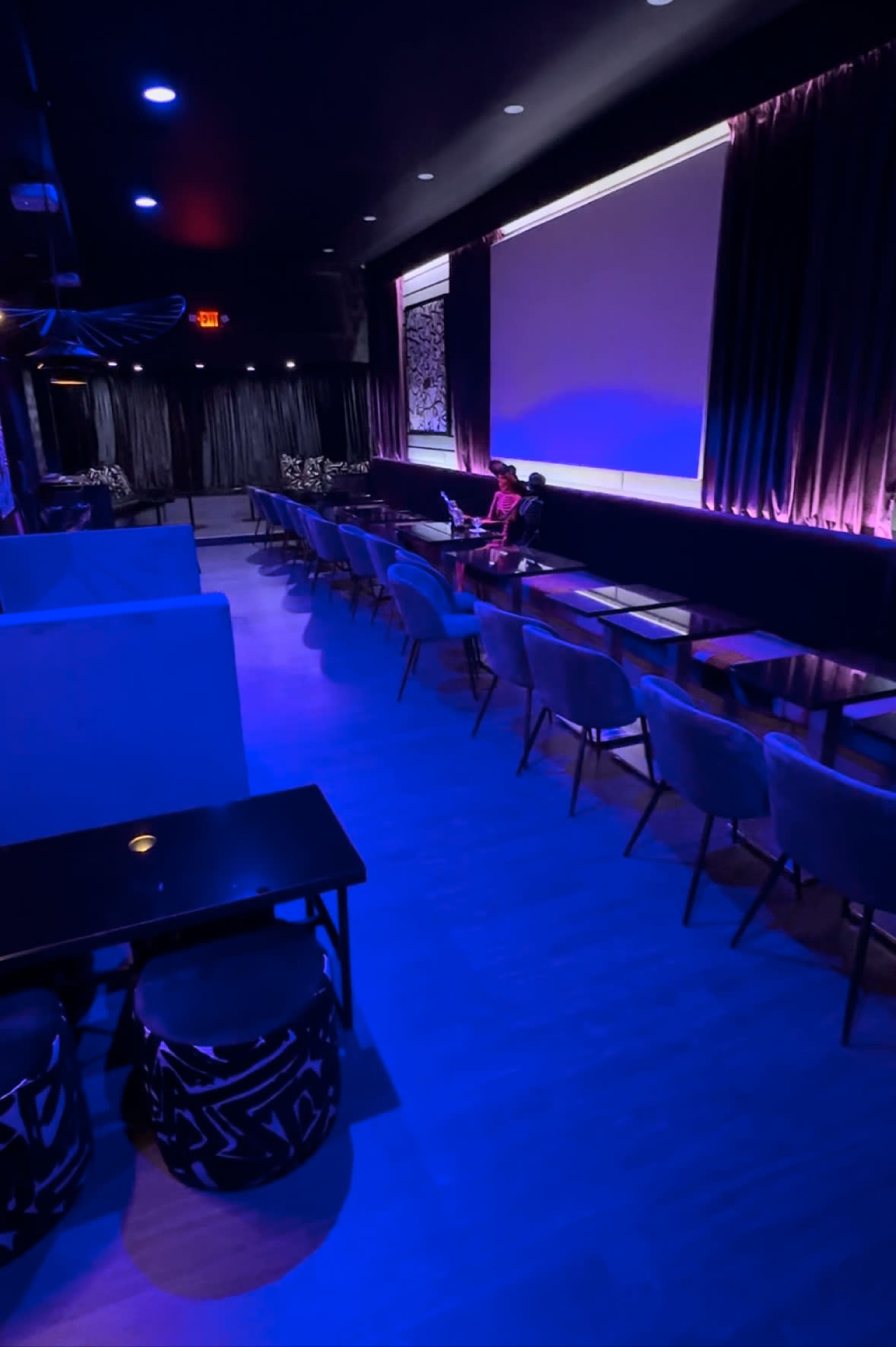 A dimly lit lounge features rows of chairs and tables with a performer seated on stage against a backdrop of curtains and ambient lighting.