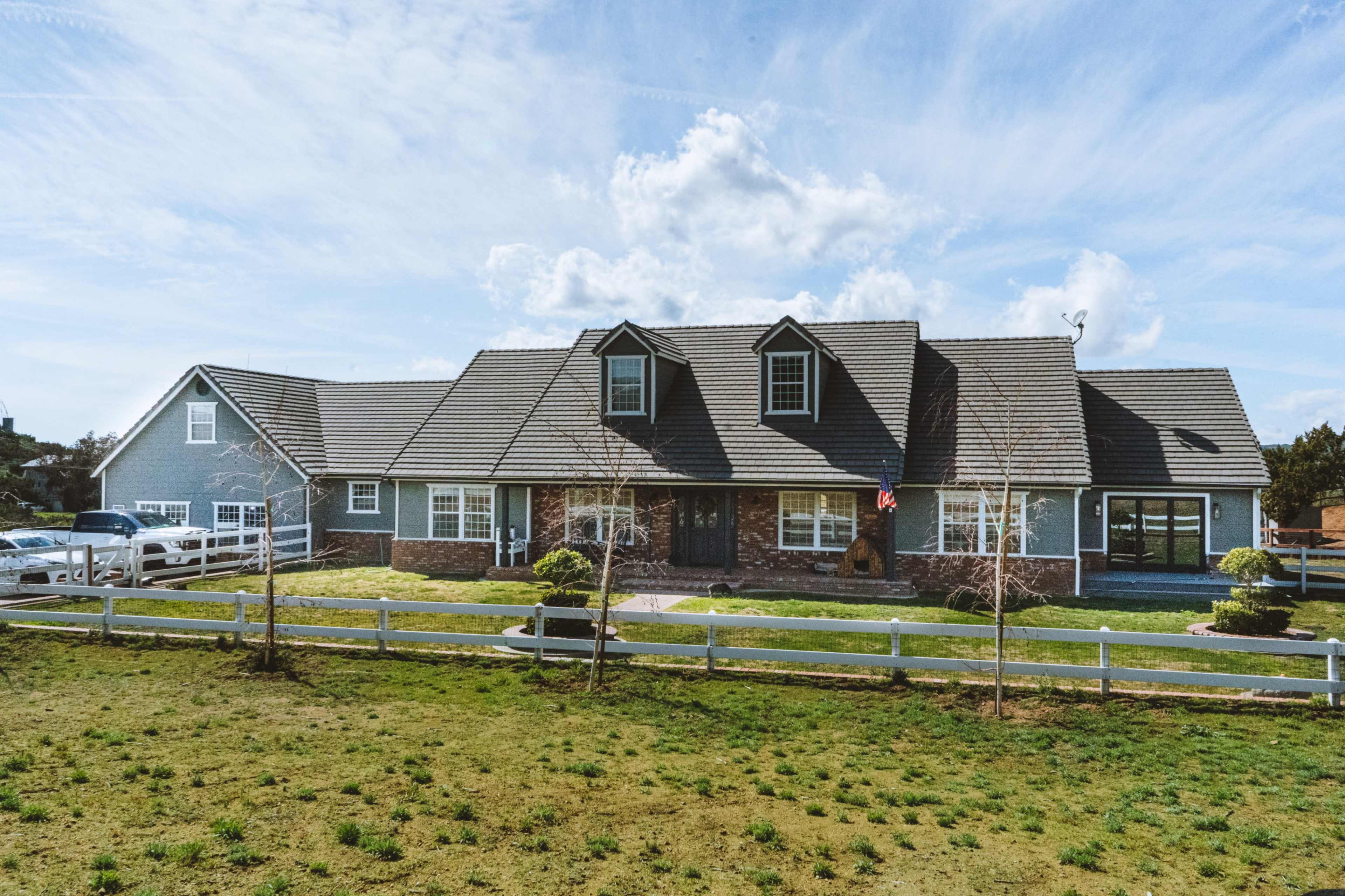 A large, two-story, blue house with a gray roof and a front lawn bordered by a white fence is shown under a clear sky.