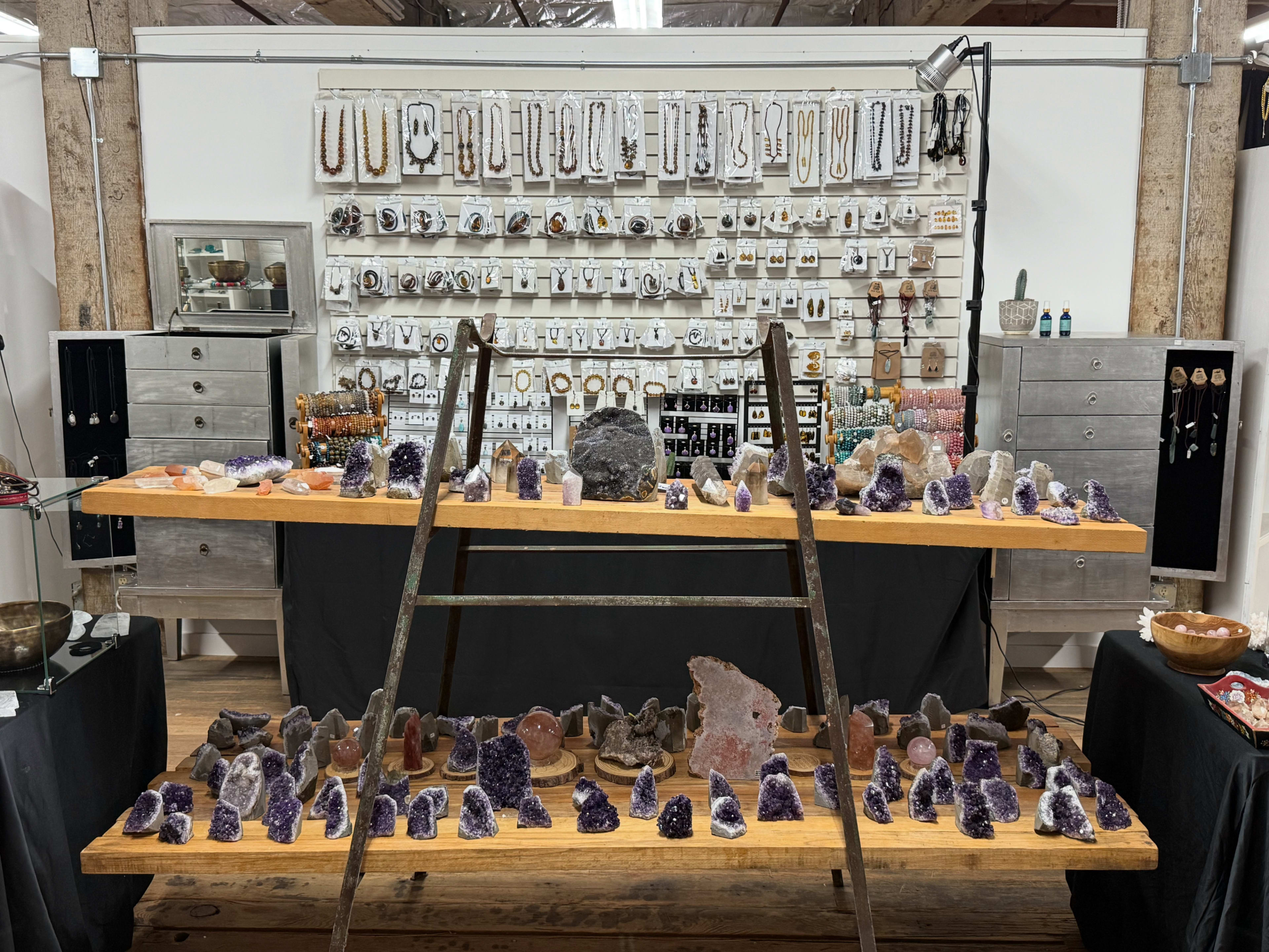 A display of various crystals and jewelry is arranged on wooden tables in a shop, with a background of hanging jewelry and other decorative items.