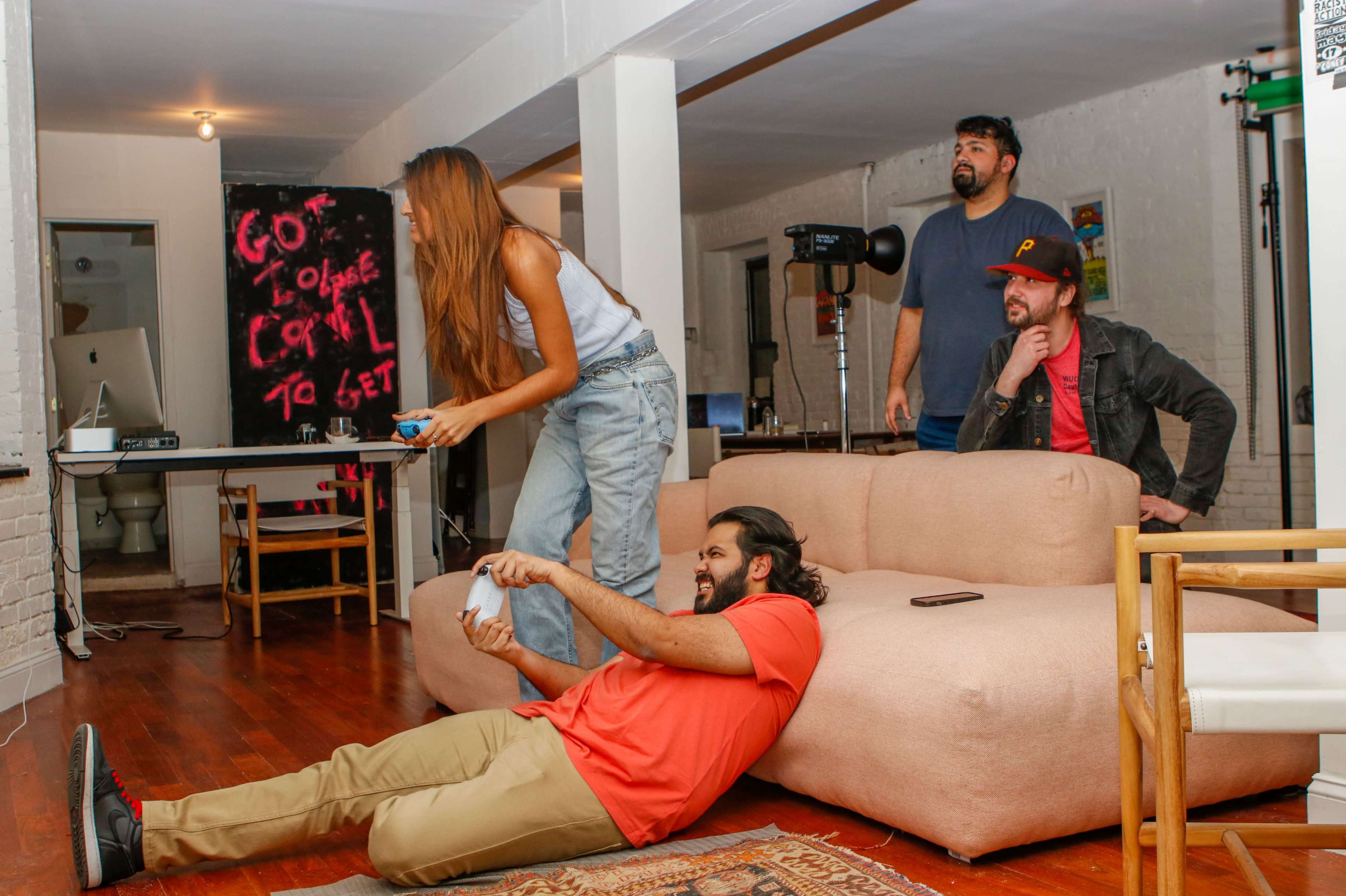 A group of four people is gathered in a modern living space, with one person lying on the floor playing a video game while the others watch from a couch and a nearby chair.