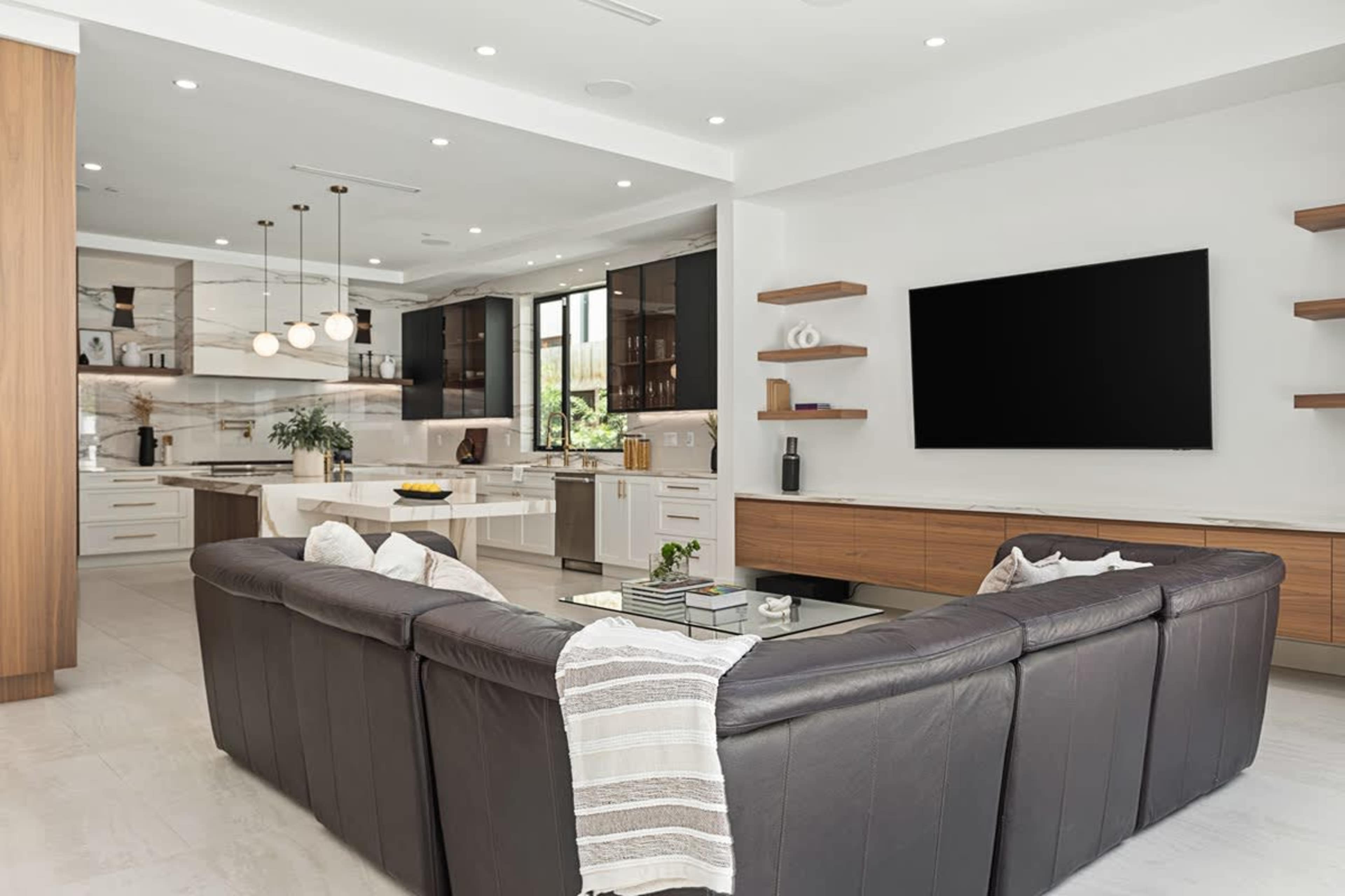 A modern living room with a sectional sofa, a coffee table, and a flat-screen television mounted on the wall, adjacent to an open kitchen with sleek cabinetry.