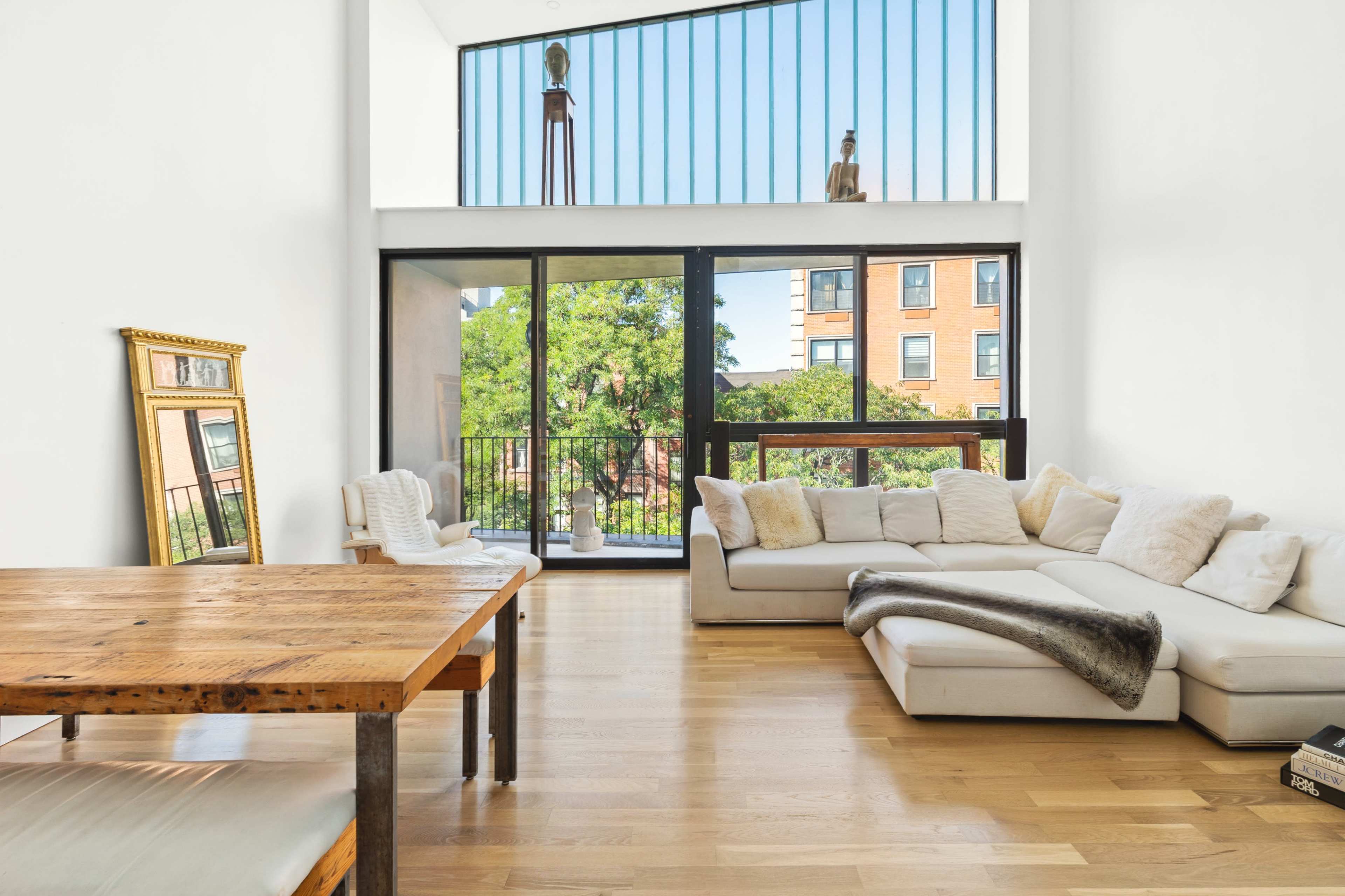 The image depicts a modern living room with a light-colored sofa, a wooden dining table, and large windows that provide a view of greenery outside.