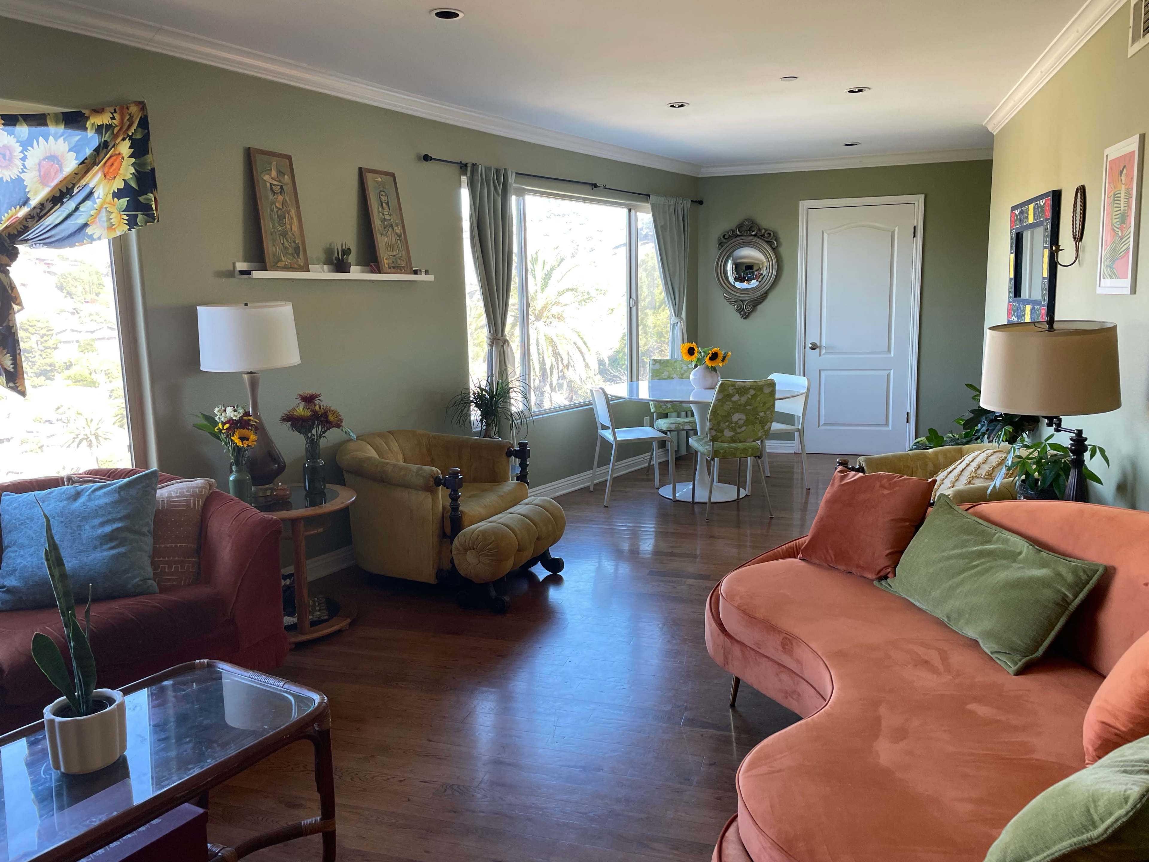 The living room features a mix of orange and green furniture, a dining area with a circular table, and large windows letting in natural light.