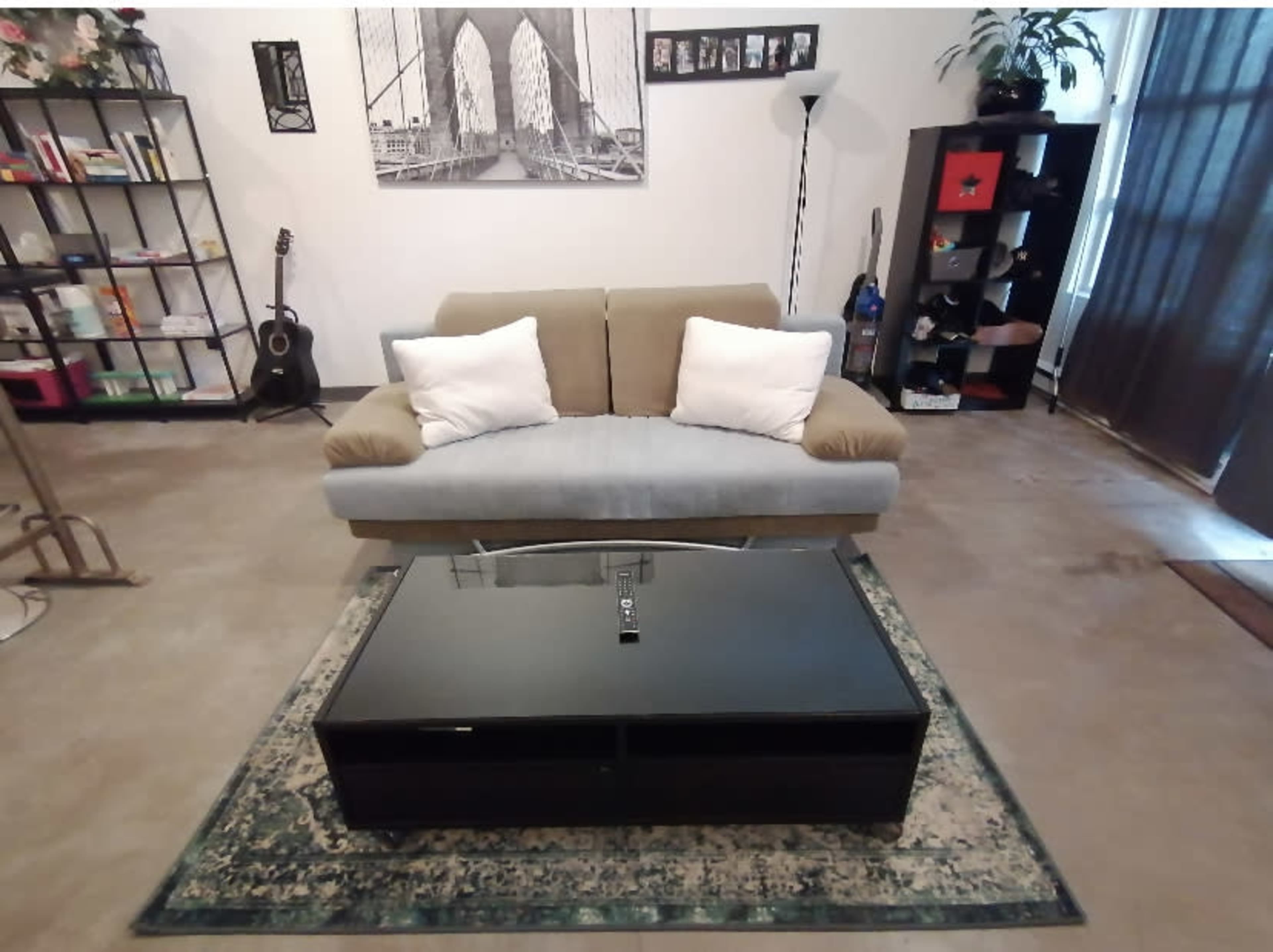 A living room with a two-tone couch, a black coffee table on an area rug, bookshelves with various items, and a guitar leaning against one of the shelves.