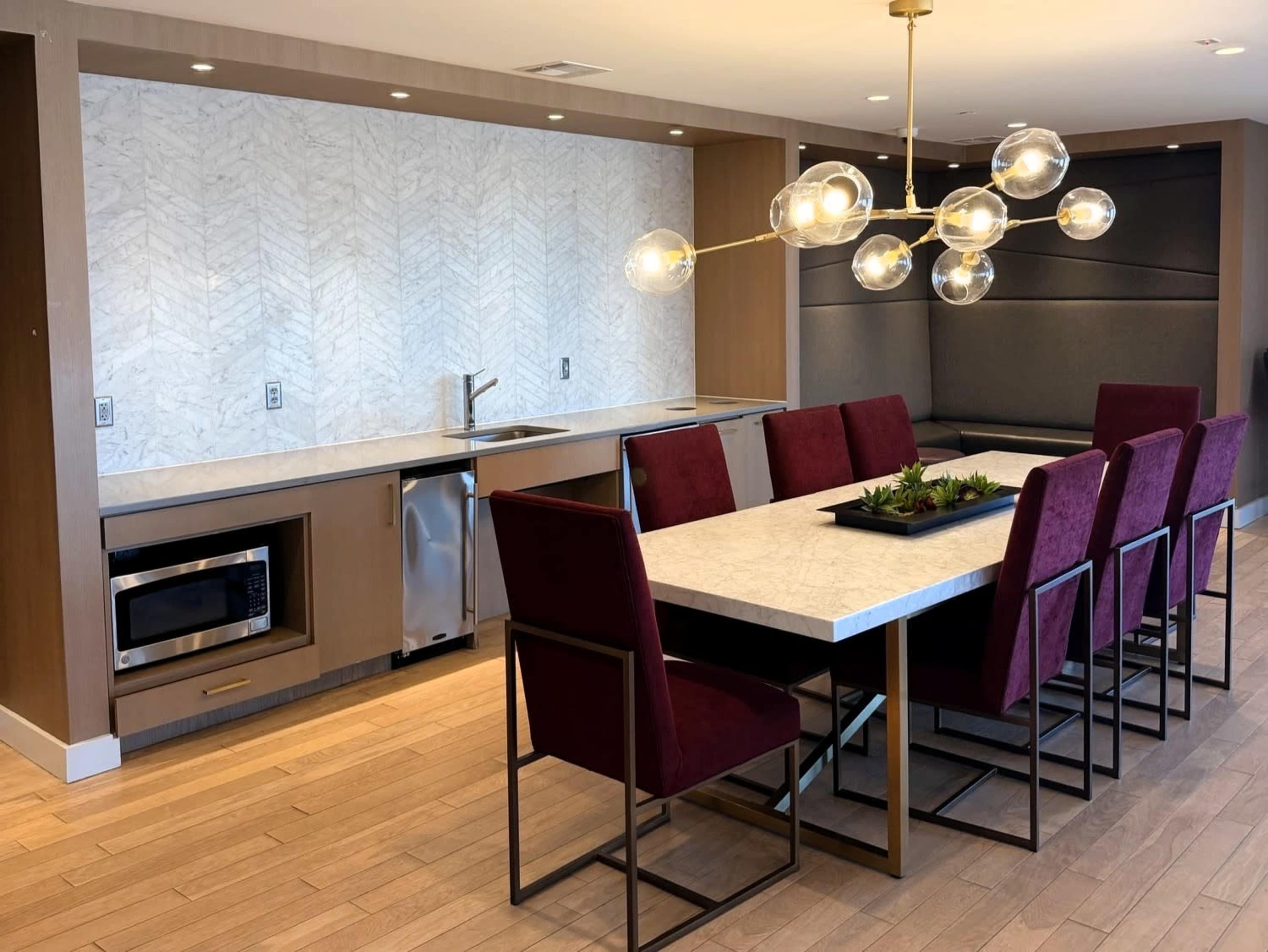 The image shows a modern kitchen and dining area featuring a long table with eight burgundy chairs, a marble countertop, and a decorative light fixture above.