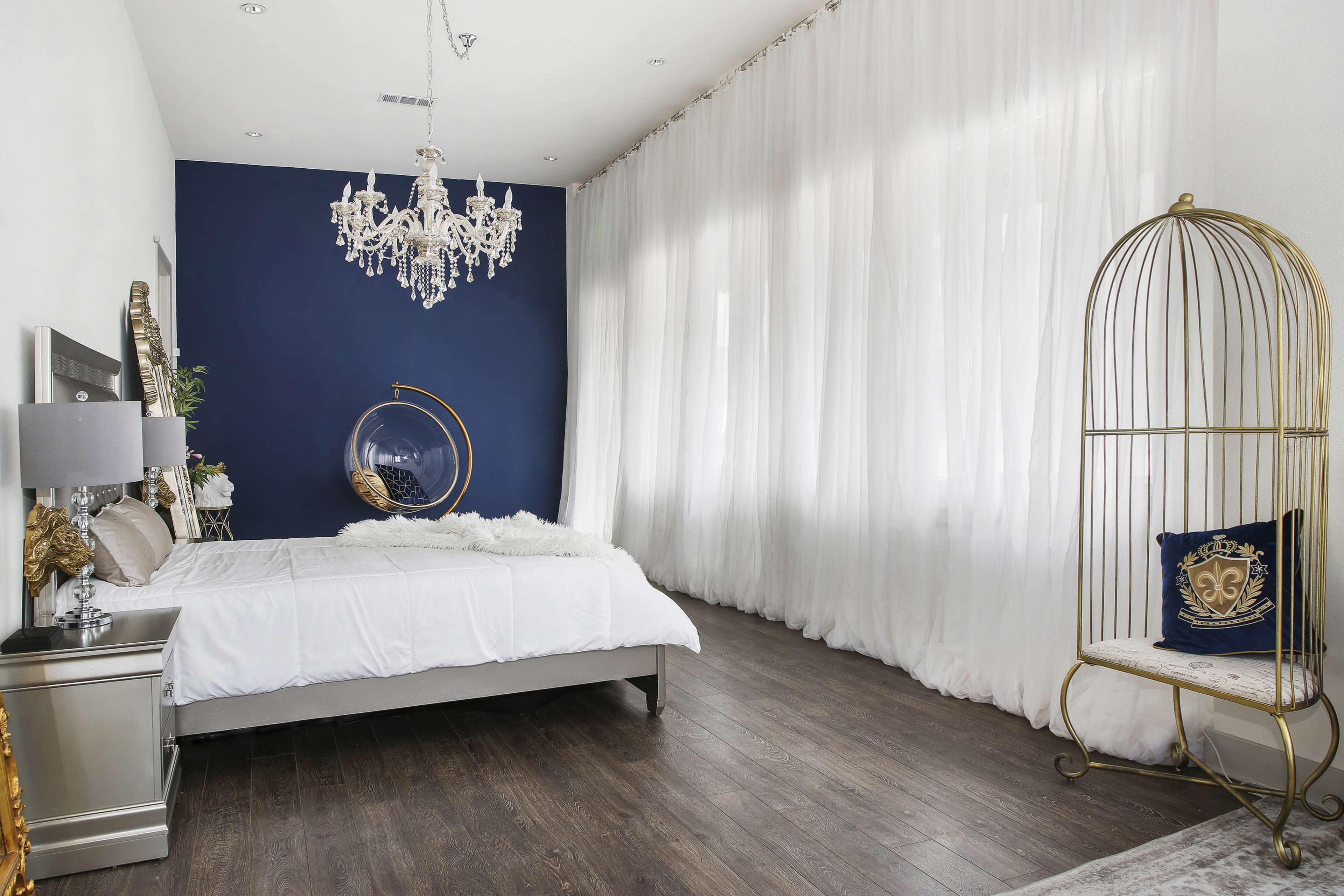 The image shows a modern bedroom featuring a bed with white linens, a large chandelier, and a decorative gold birdcage beside a window draped with sheer white curtains.
