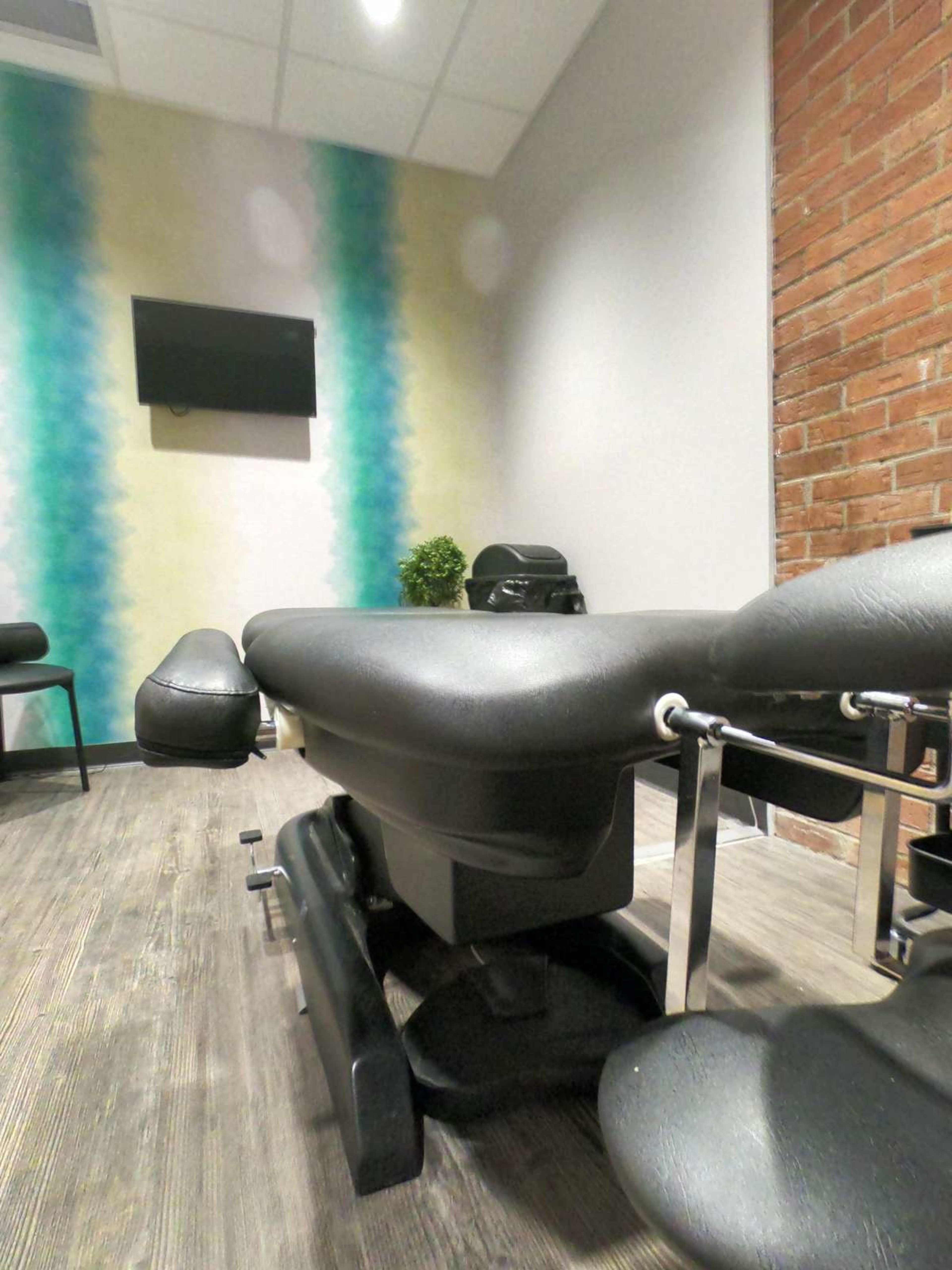 The image shows a treatment room with a black massage table, a small potted plant, and a television on a light-colored wall with a gradient design.